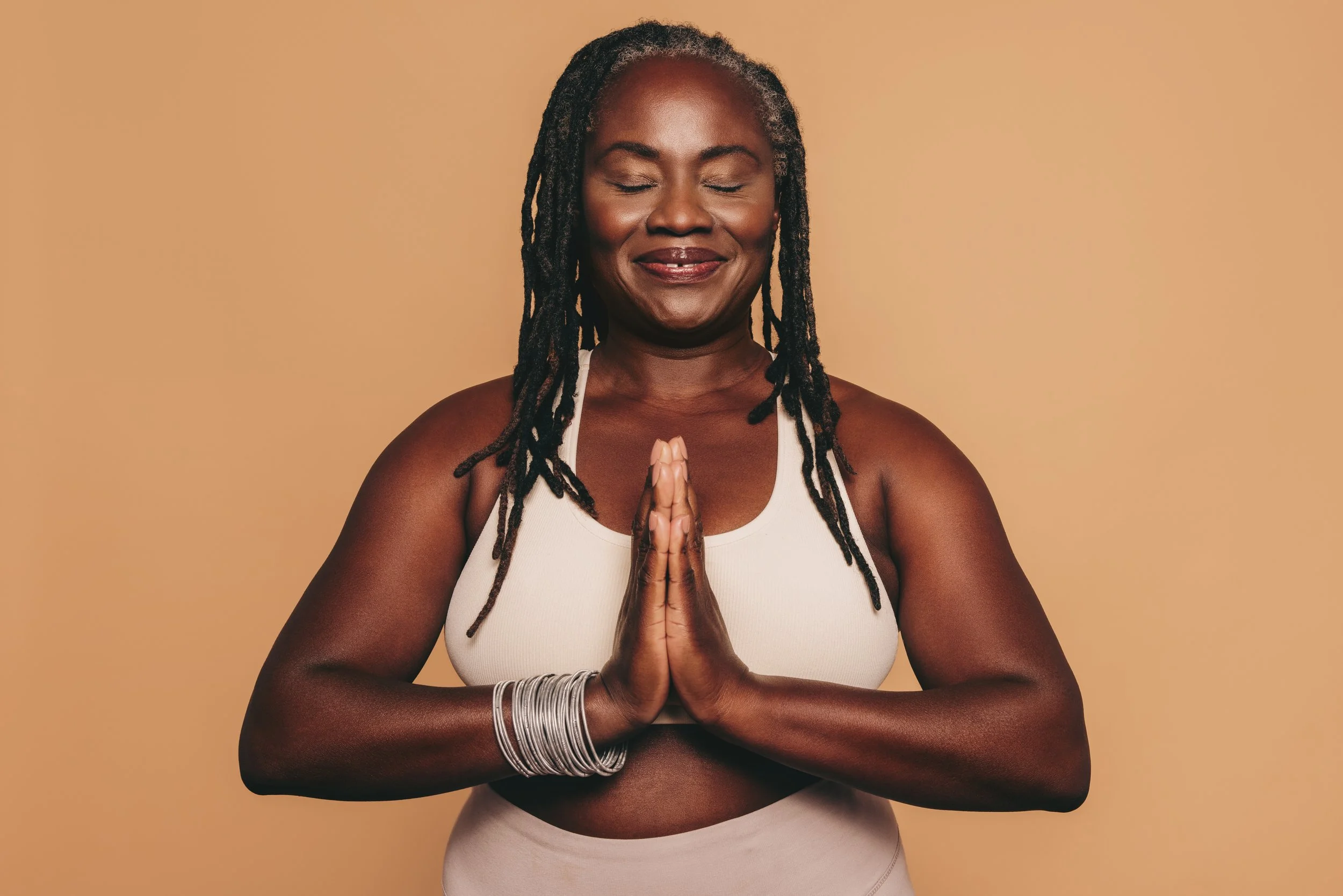 A woman with dreadlocks in a white sports bra and beige pants is standing against a beige background, with her hands pressed together in a prayer position and eyes closed.