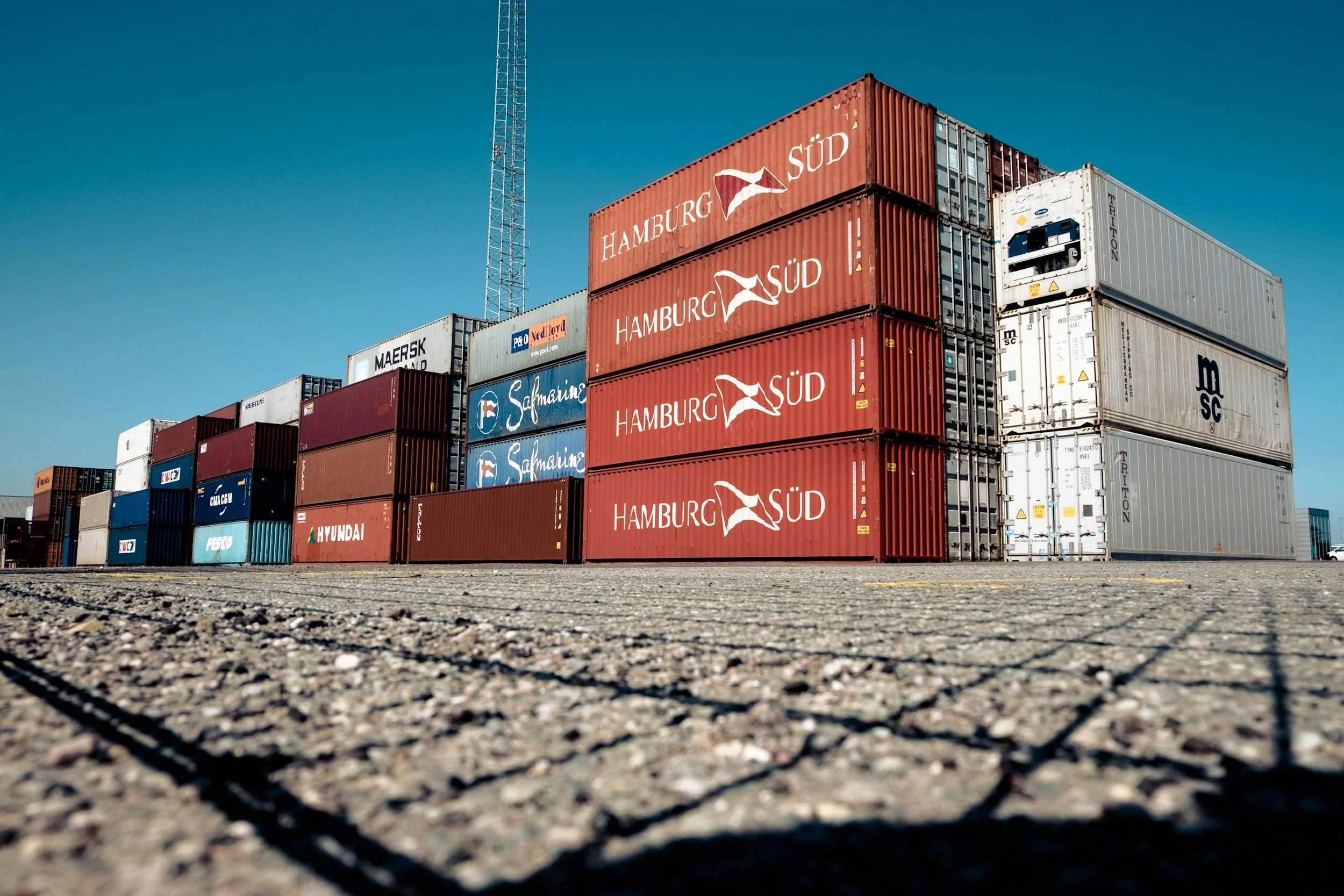 Multiple stacked shipping containers at a port with a clear blue sky in the background.