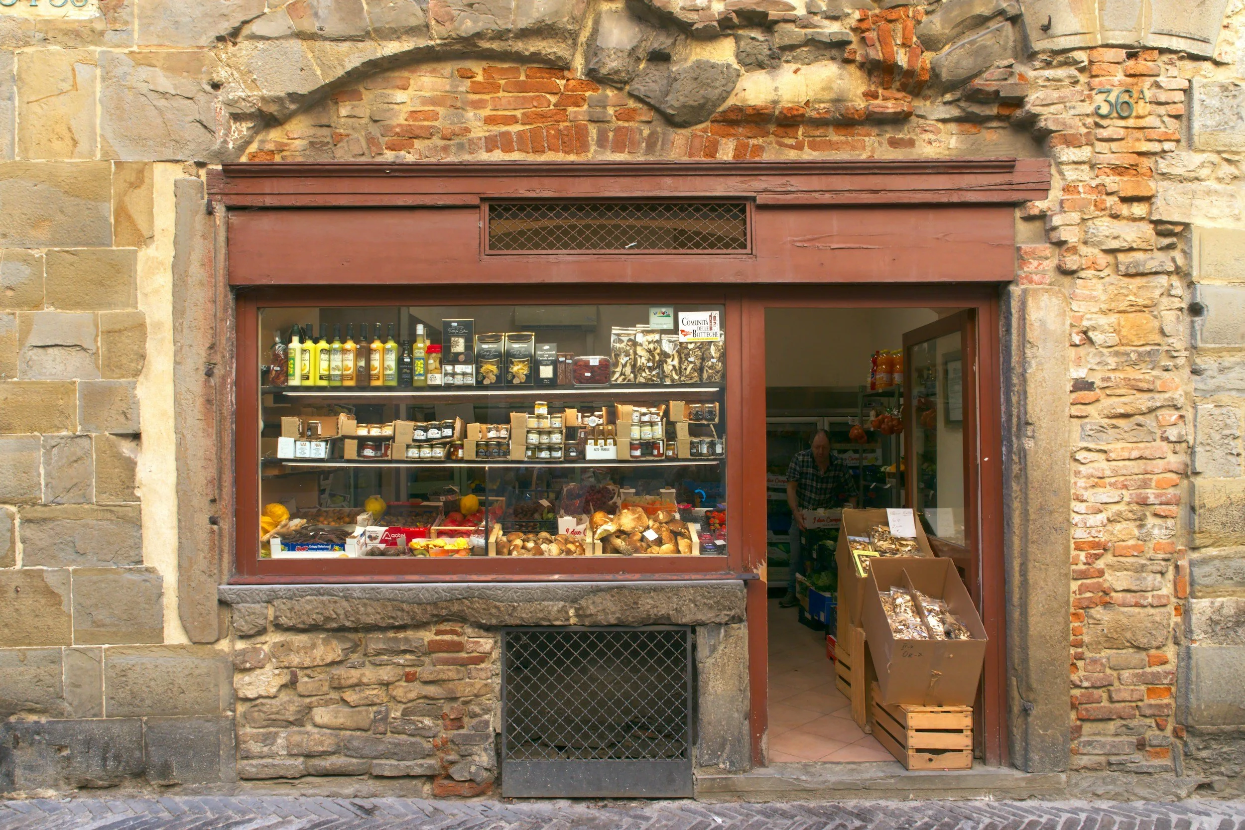 A small shop with a stone and brick facade, displaying produce and jars through the window, and a man working inside.
