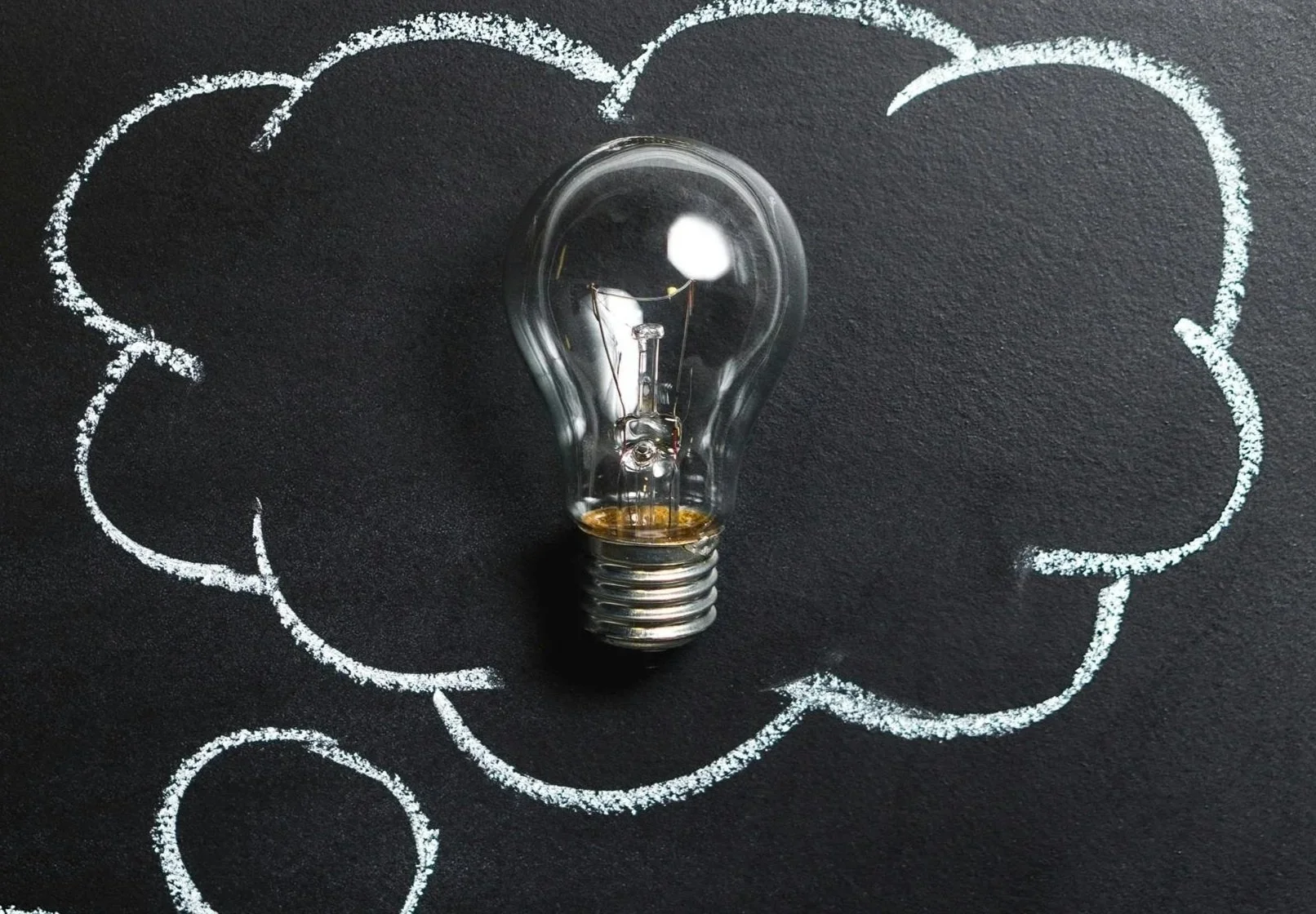 A clear light bulb placed on a black surface with white chalk drawing of a cloud and a thought bubble around it.