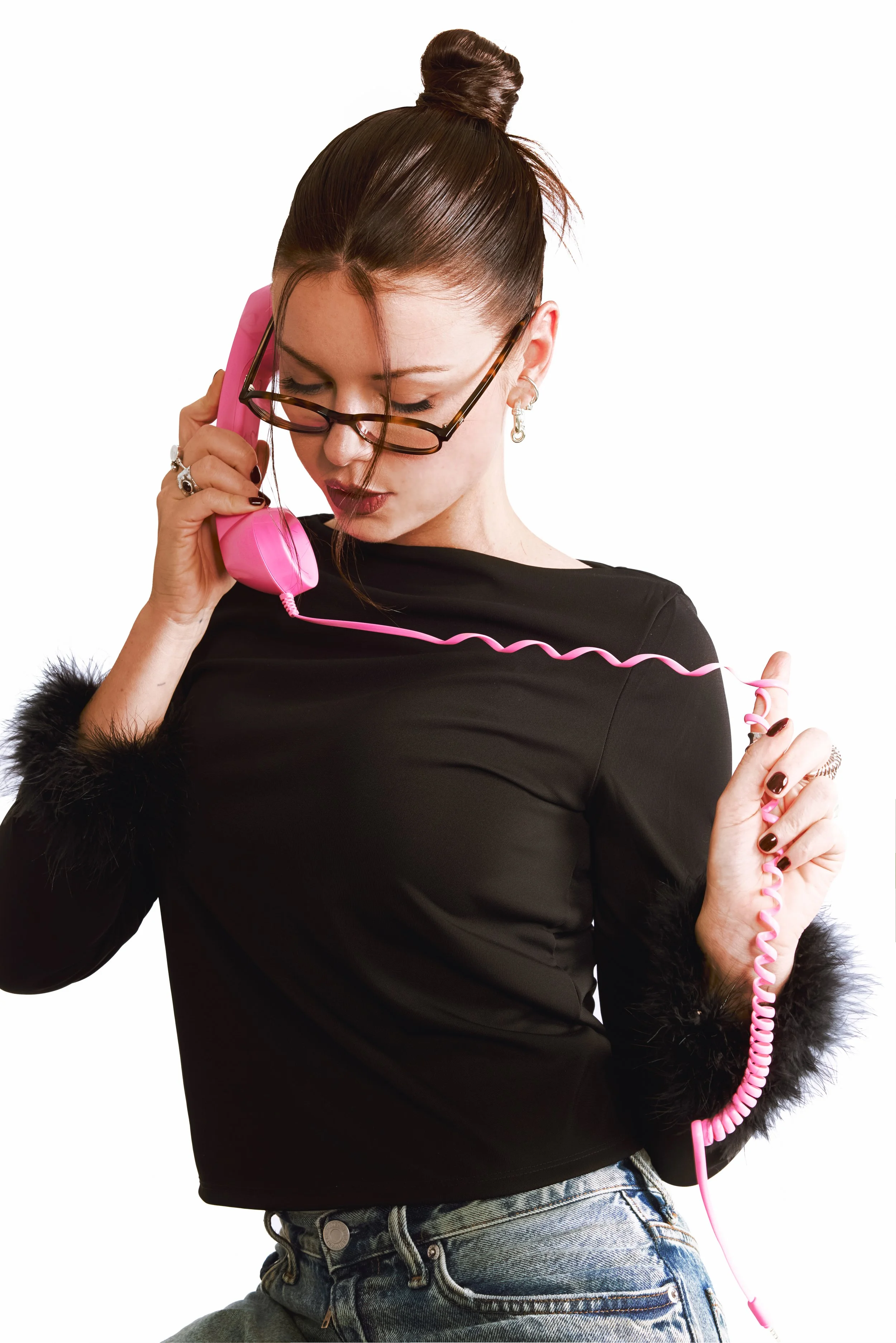 Félice Bricca, co-fondatrice de Care Care Agency avec des lunettes, portant un pull noir avec des poignets en pompons noirs, tenant un téléphone rose ancien et portant une bague en argent, regardant vers le bas.