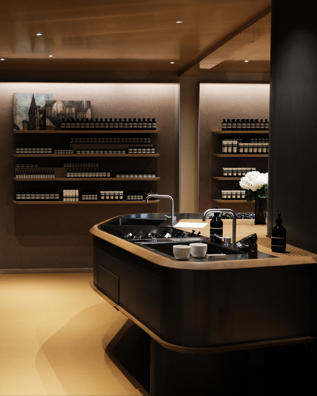 Un espace de préparation Aesop avec un comptoir noir en bois clair, un évier avec robinet, des fleurs blanches en vase, des lotions ou savons, et des étagères en arrière-plan remplies de petits contenants identiques.