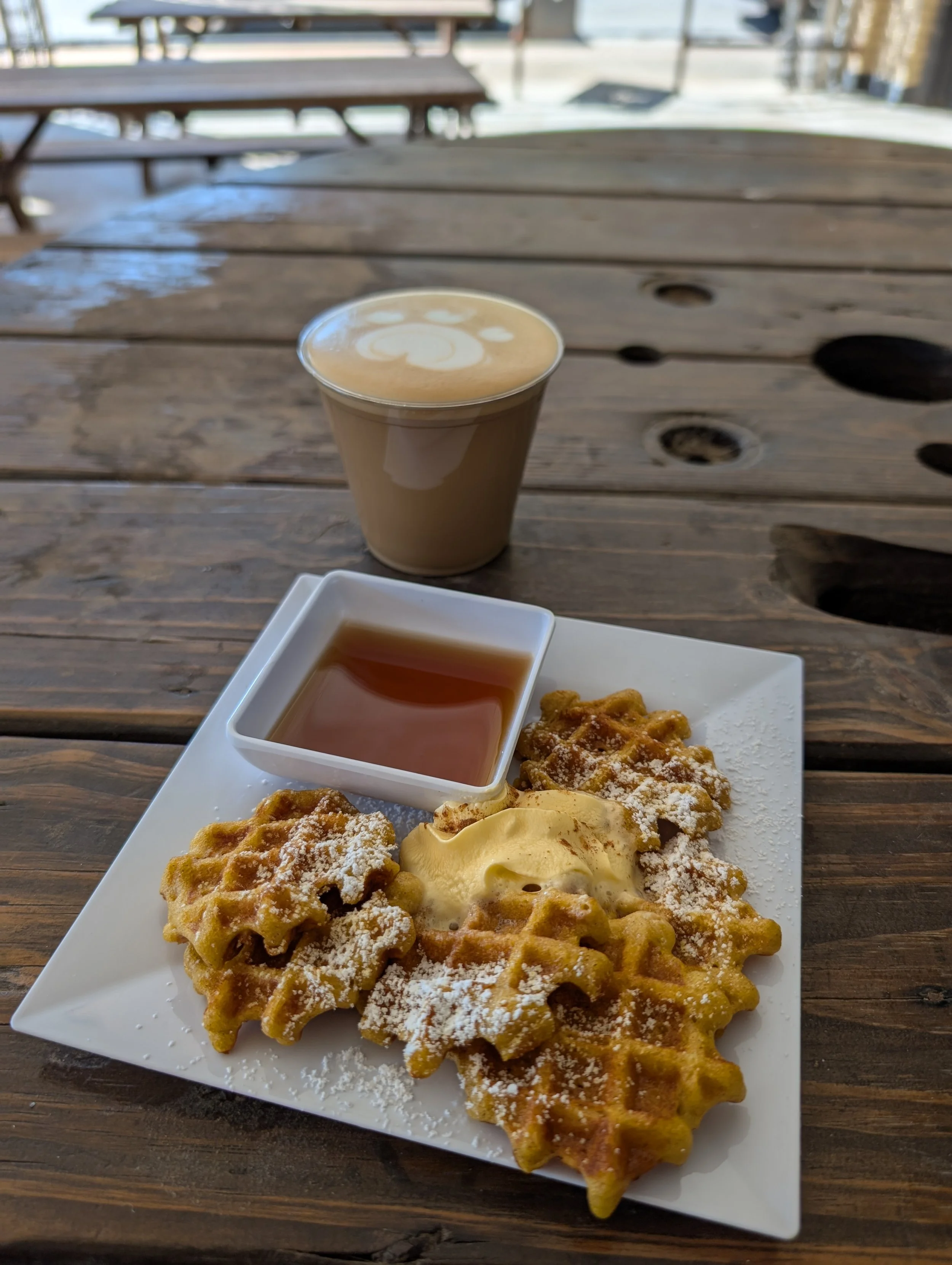 A plate of waffles topped with powdered sugar and butter, served with syrup in a small square dish, accompanied by a latte in a clear plastic cup on a wooden table outdoors.