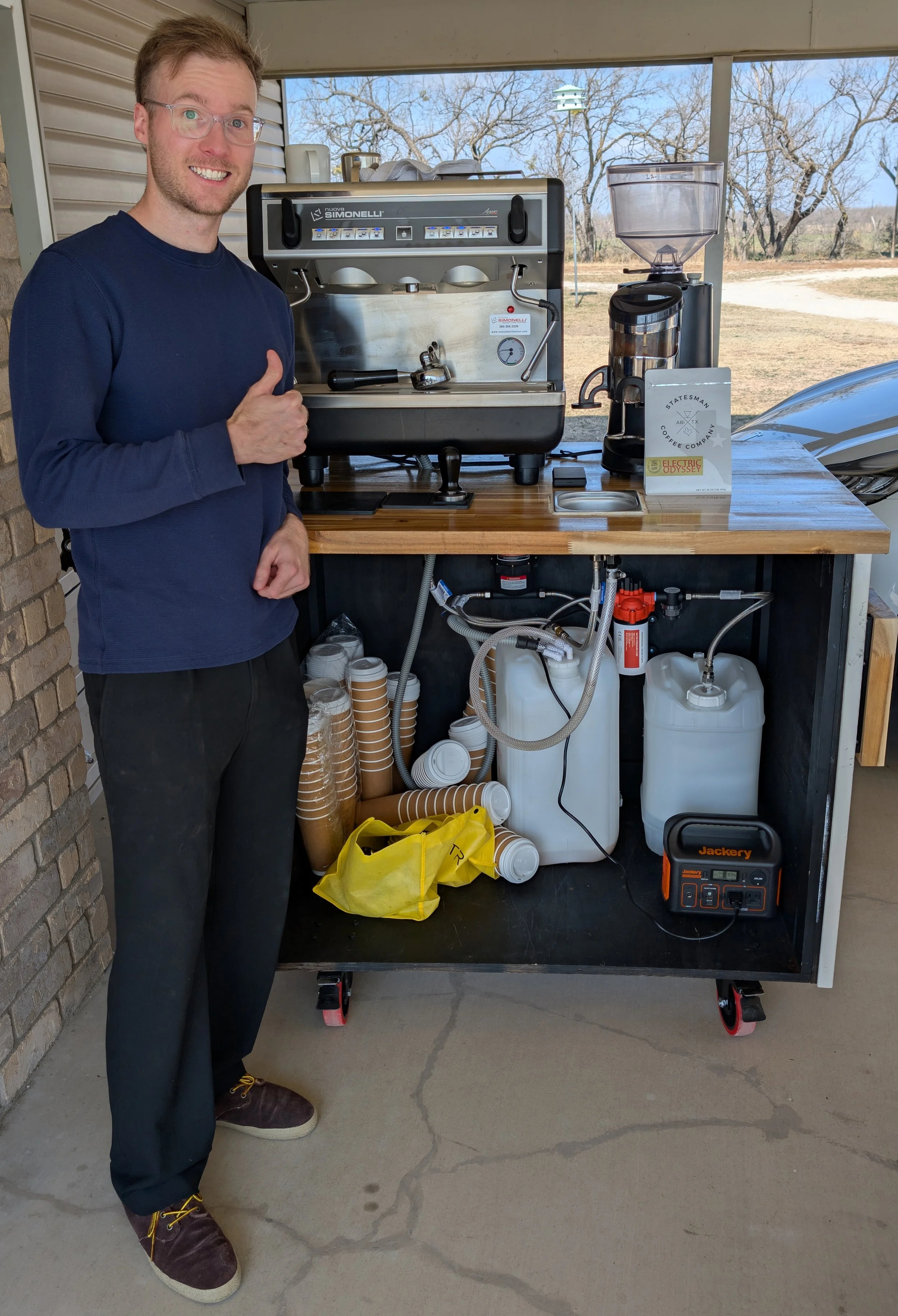 A man standing next to a coffee-making station outdoors on a porch, giving a thumbs-up. The setup includes a professional espresso machine, a coffee grinder, and supplies such as cups and water containers.