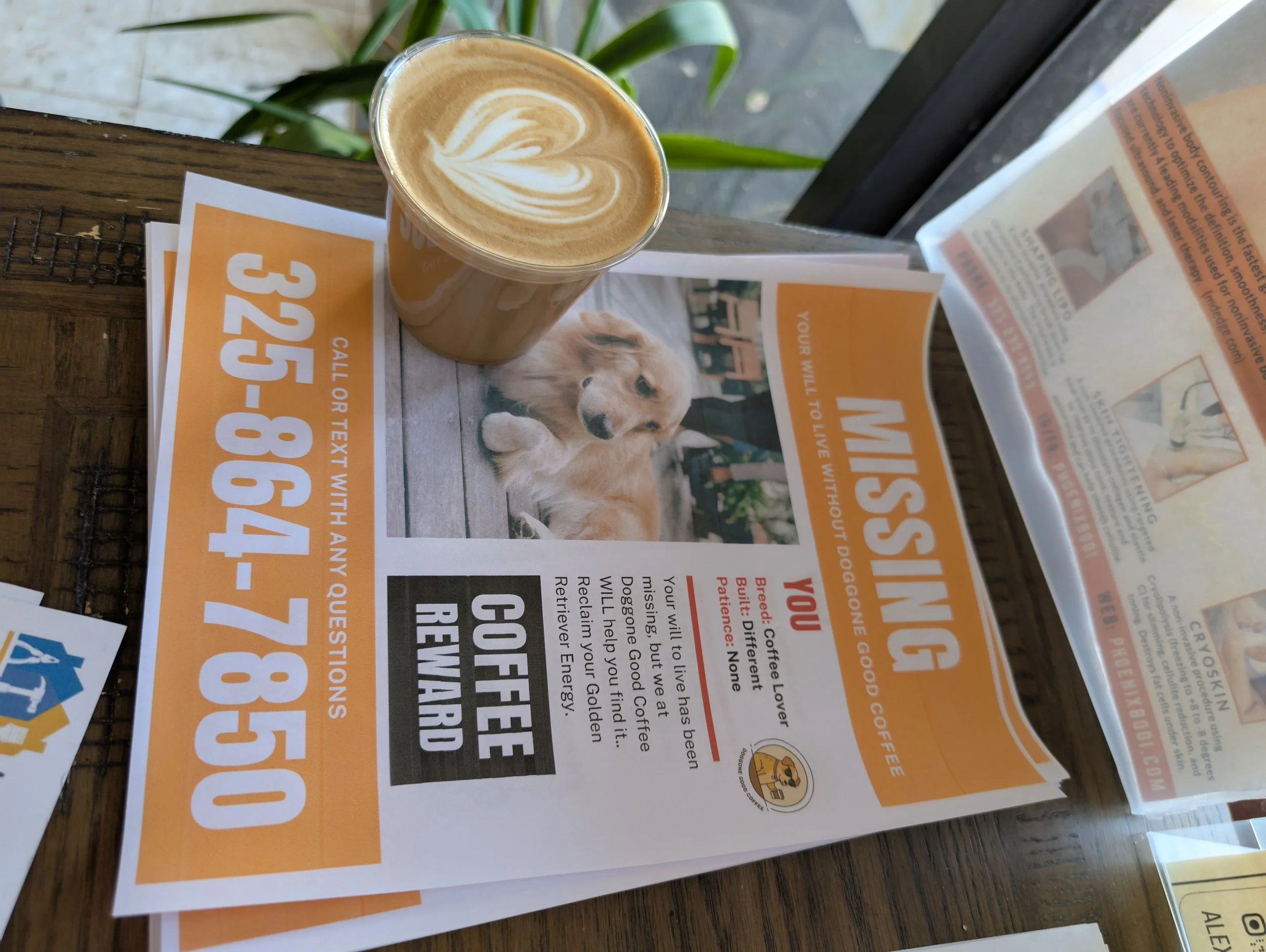 A cup of coffee with latte art on a wooden table next to parked cars and a window with a newspaper on it.