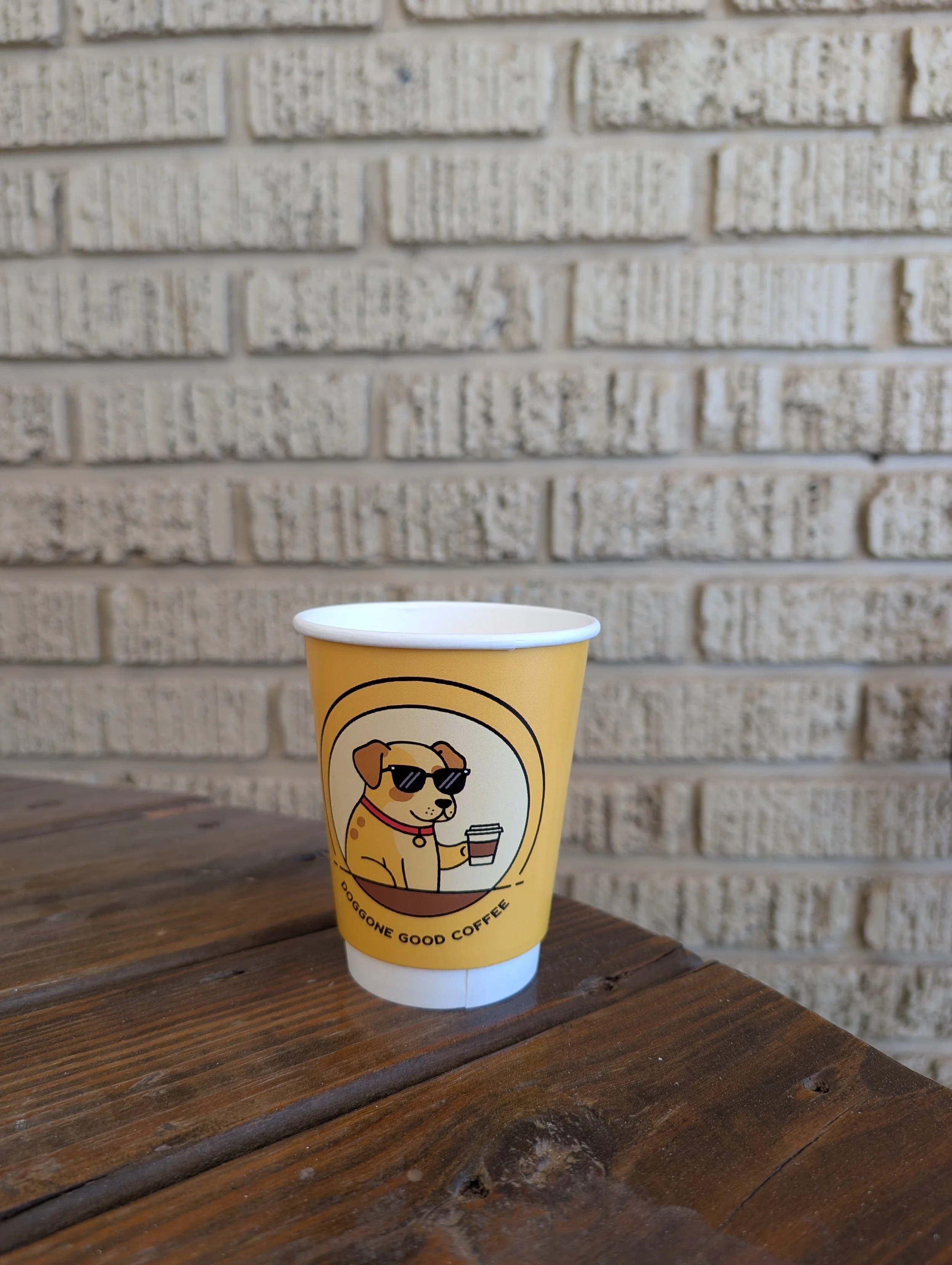 A yellow coffee cup with a cartoon dog wearing sunglasses and holding a coffee cup, sitting on a wooden surface against a brick wall background.