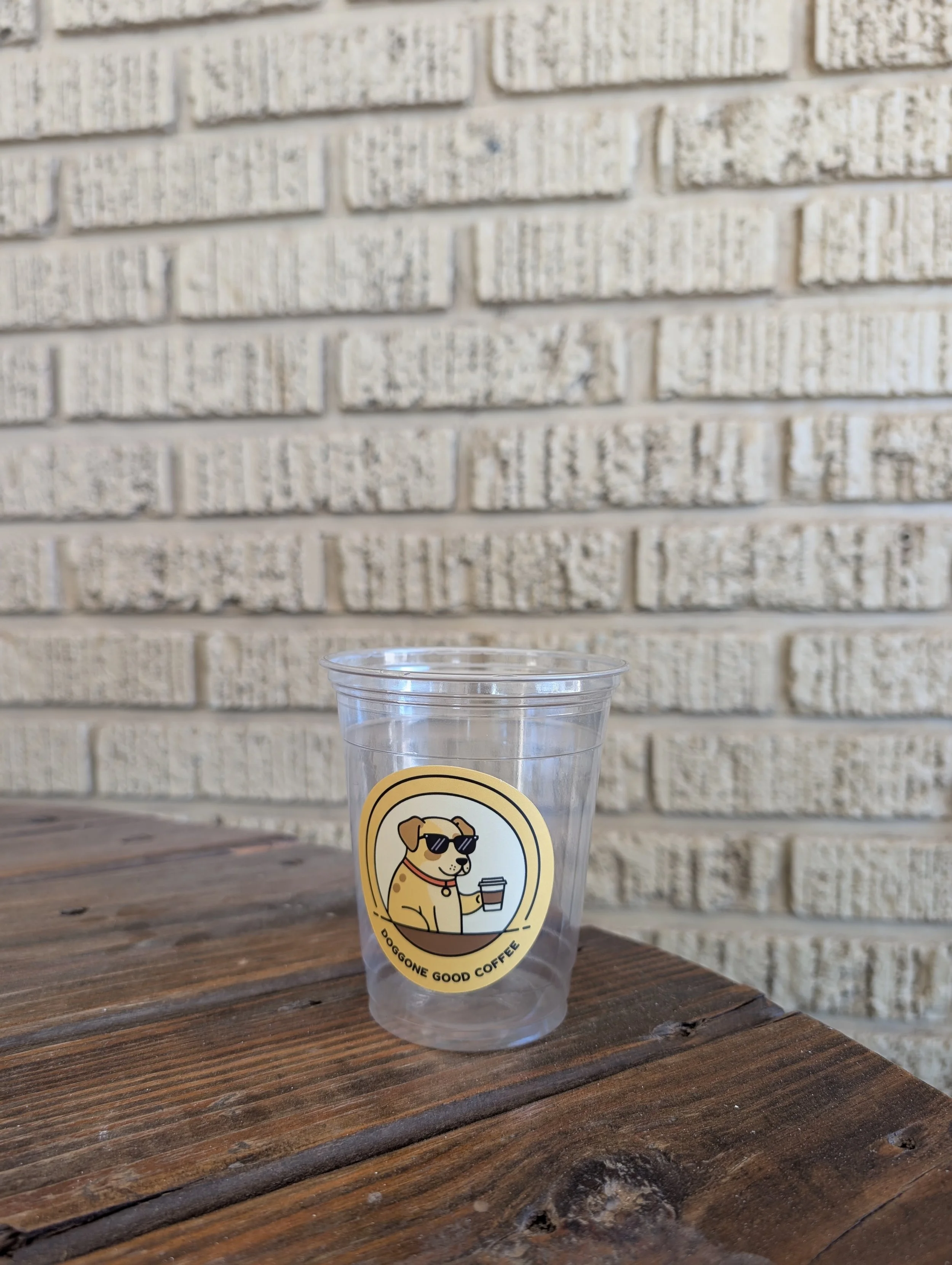 A clear plastic cup with a sticker of a dog wearing sunglasses and holding a coffee cup, with text reading 'Doggone Good Coffee,' placed on a wooden surface against a beige brick wall.