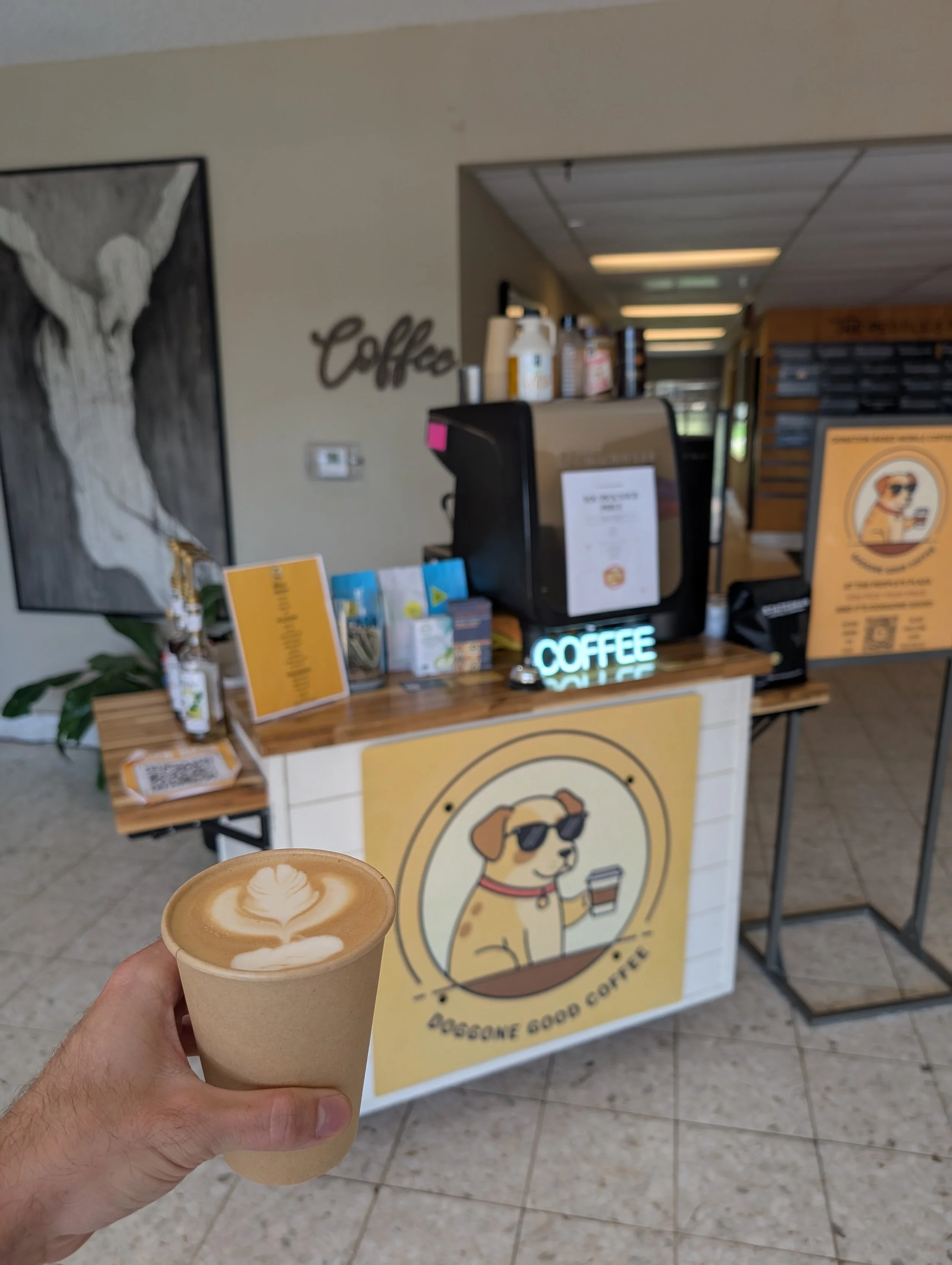 A person holding a cup of coffee with latte art in front of a coffee stand with a dog logo, inside a cafe or coffee shop.