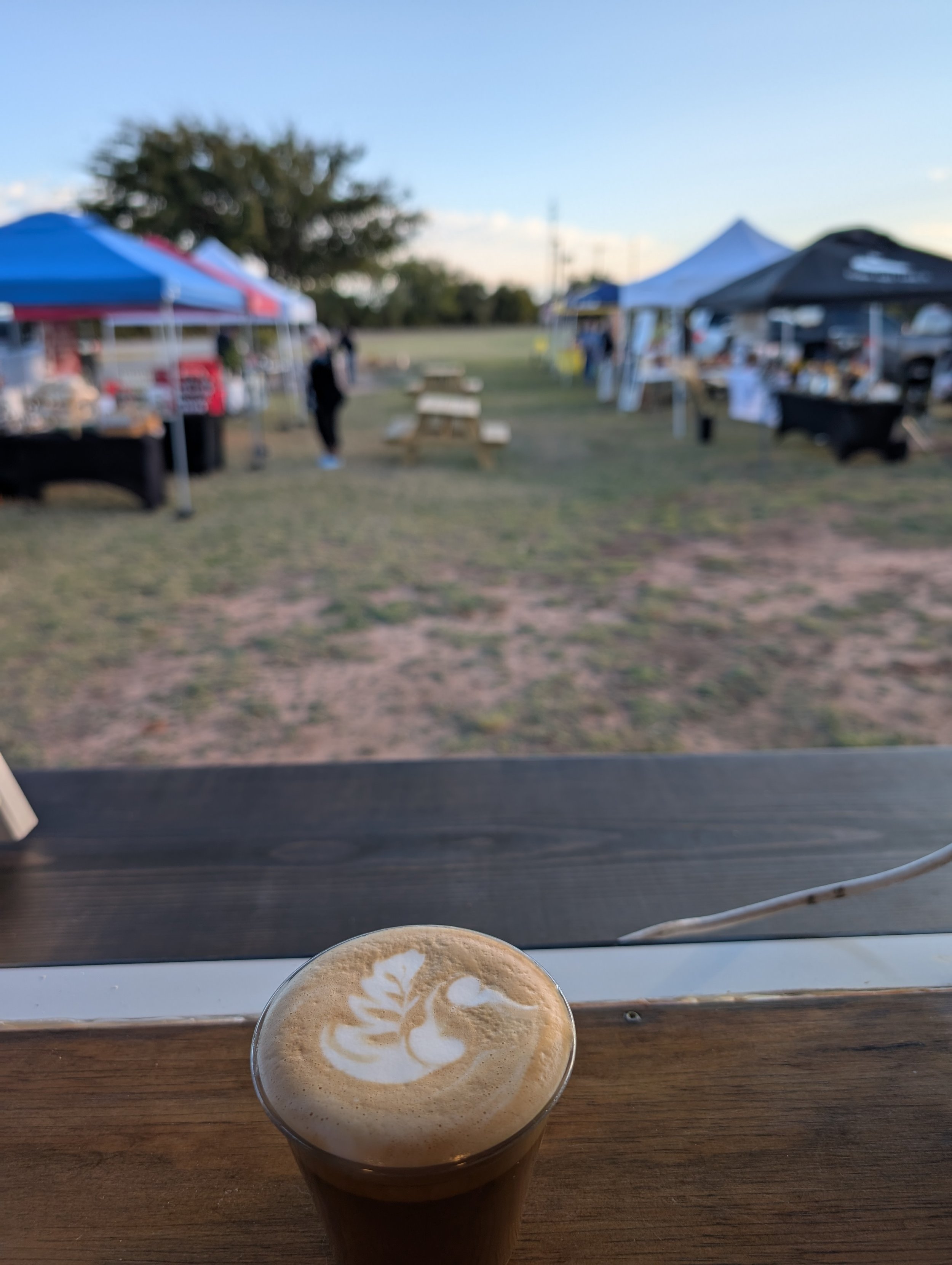 A coffee with latte art on top, viewed from inside a cafe, with outdoor market tents and people in the background.