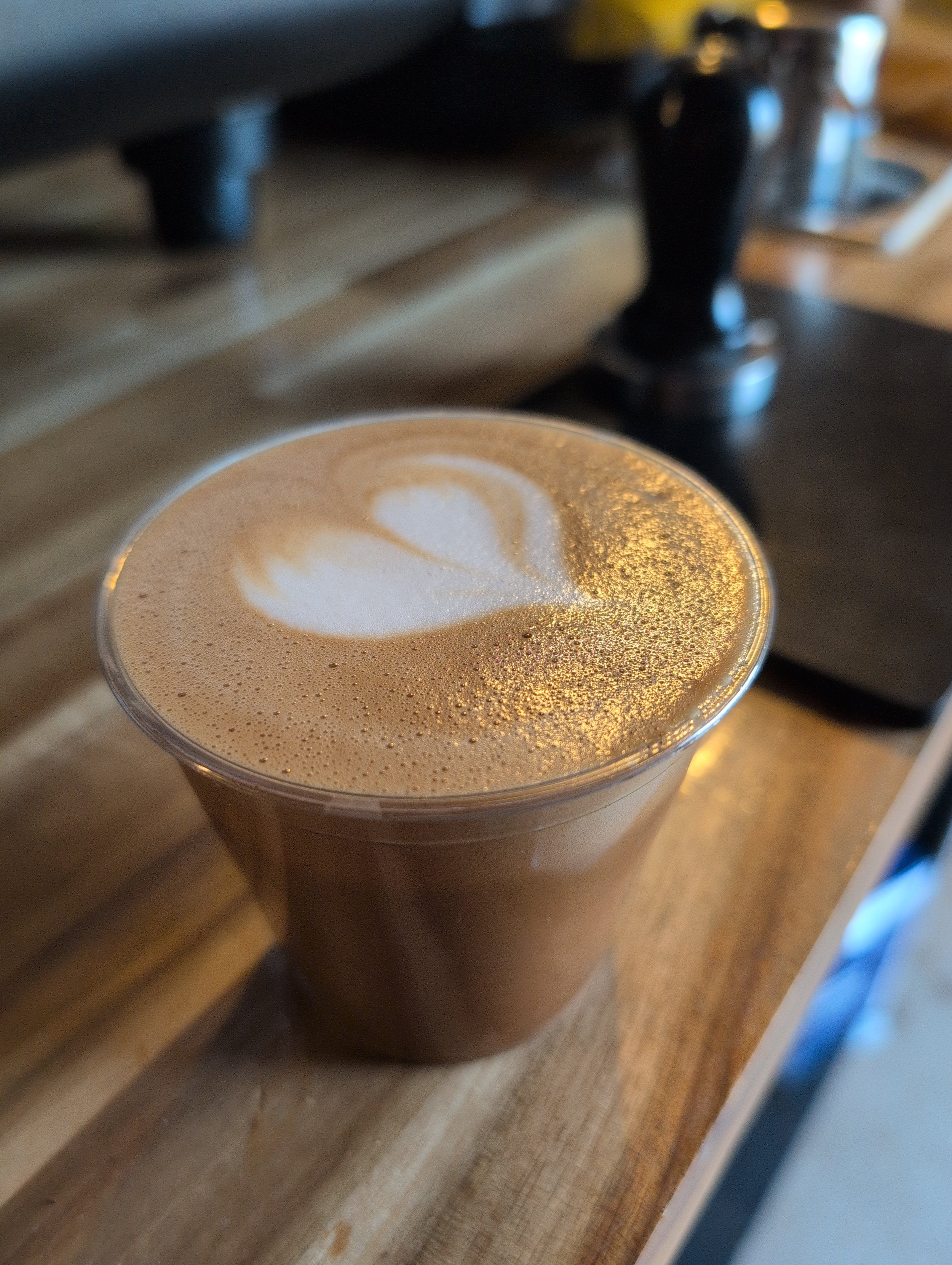 A close-up of a latte in a clear plastic cup on a wooden table, with latte art in the shape of a heart and foam dusted with gold shimmer.