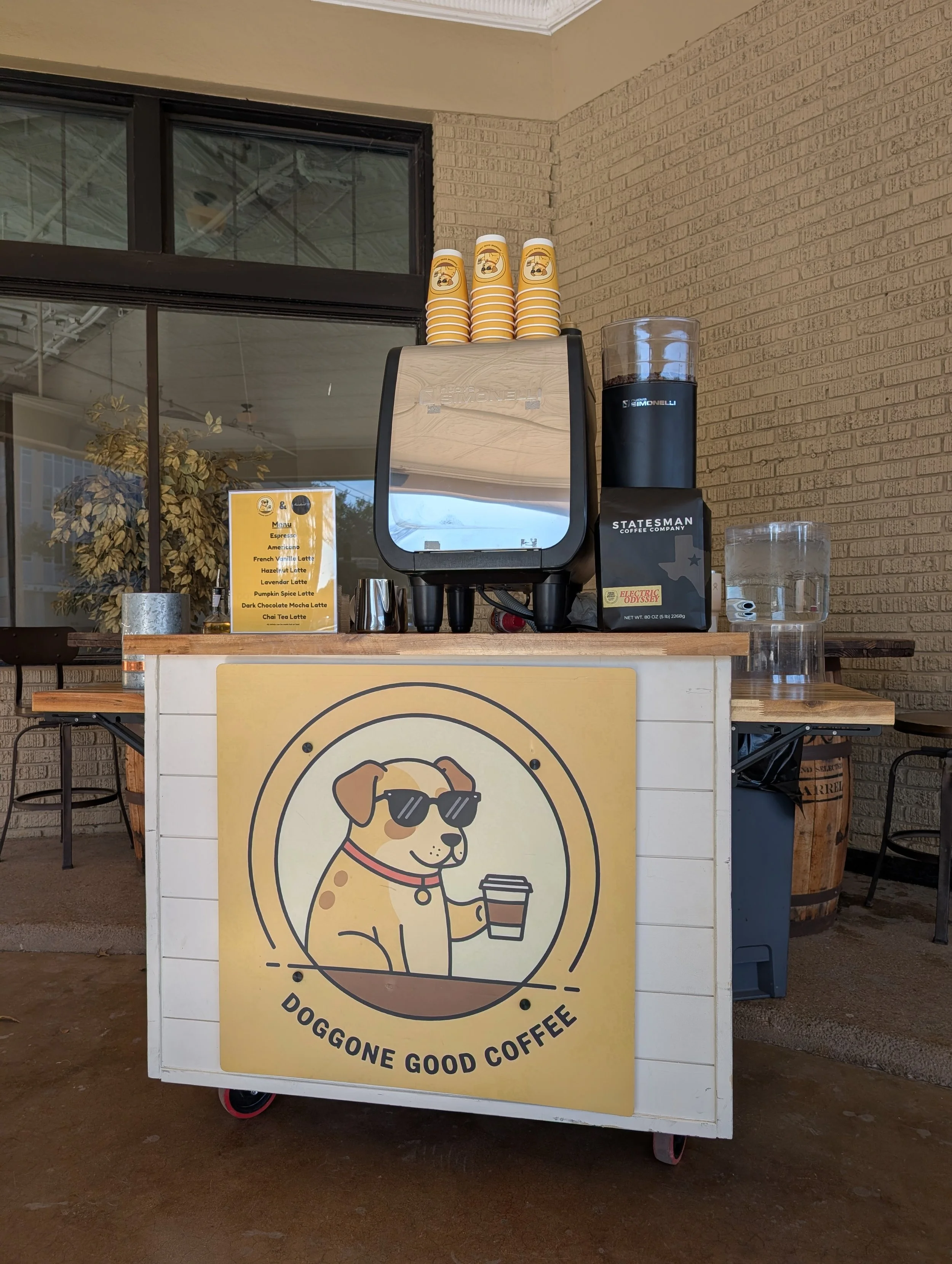 Coffee cart with a sign featuring a cartoon dog wearing sunglasses and holding a coffee cup, with the text "Doggone Good Coffee." The cart has a coffee machine, cups, and water bottles on top, located indoors near a window and brick wall.