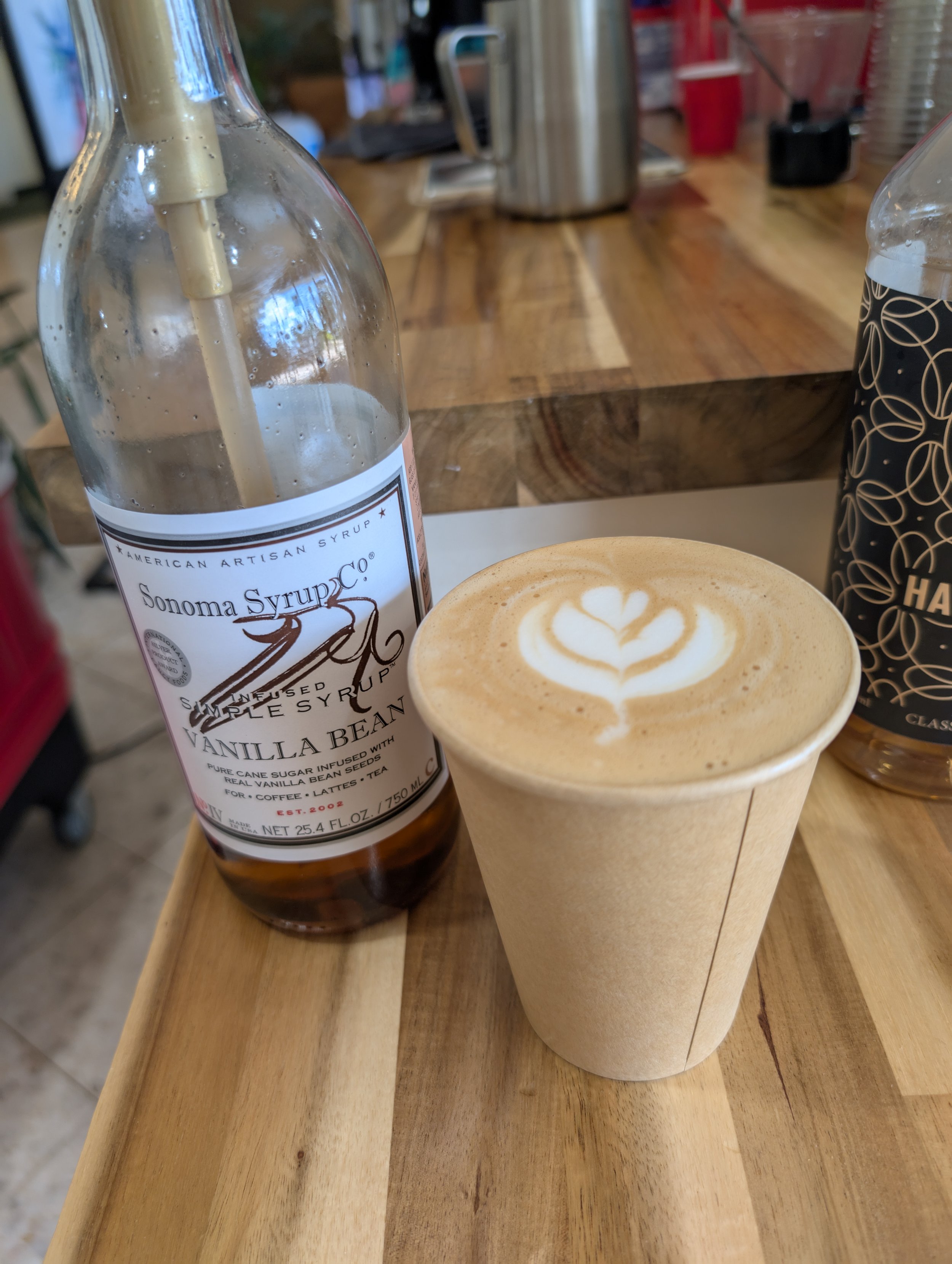 A latte with foam art in a paper cup, a bottle of Sonoma Syrup vanilla bean syrup, and a canned drink on a wooden table in a cafe.