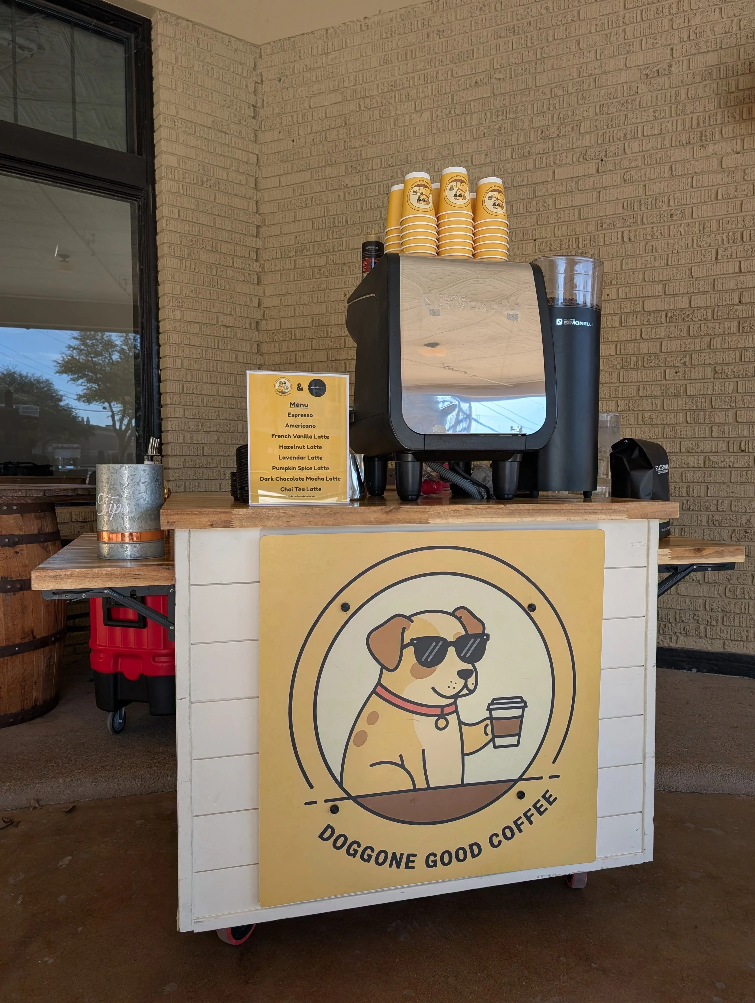 A coffee stand labeled 'Doggone Good Coffee' with a cartoon dog wearing sunglasses and holding a coffee cup, set against a brick wall. The stand has a coffee machine on top and disposable cups stacked above it. A menu sign lists different coffee drin