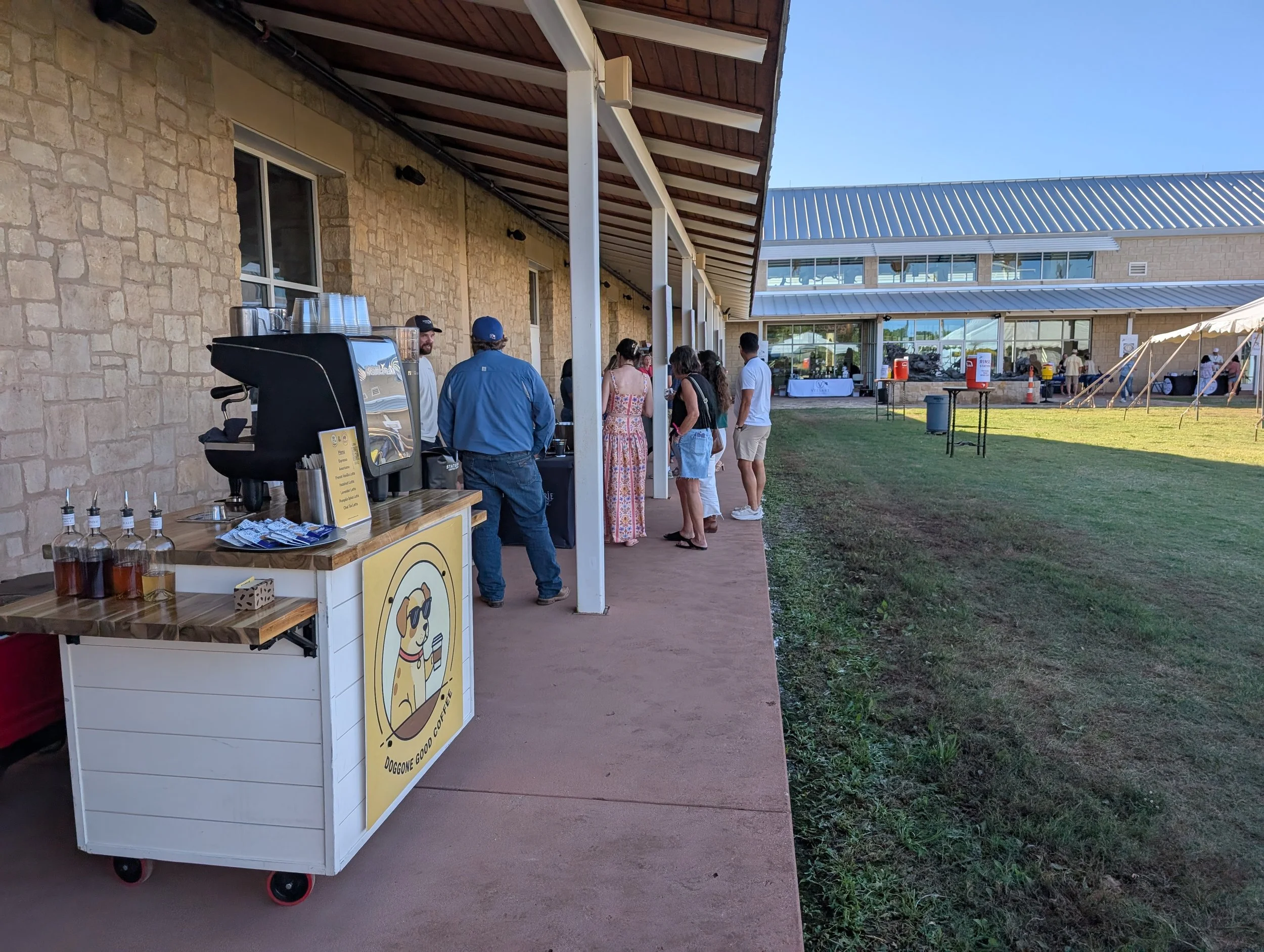People standing in line at an outdoor coffee stand, with a sign featuring a cartoon dog drinking coffee, next to a coffee machine and bottles of syrup, on a patio outside a building with a stone wall and large windows.