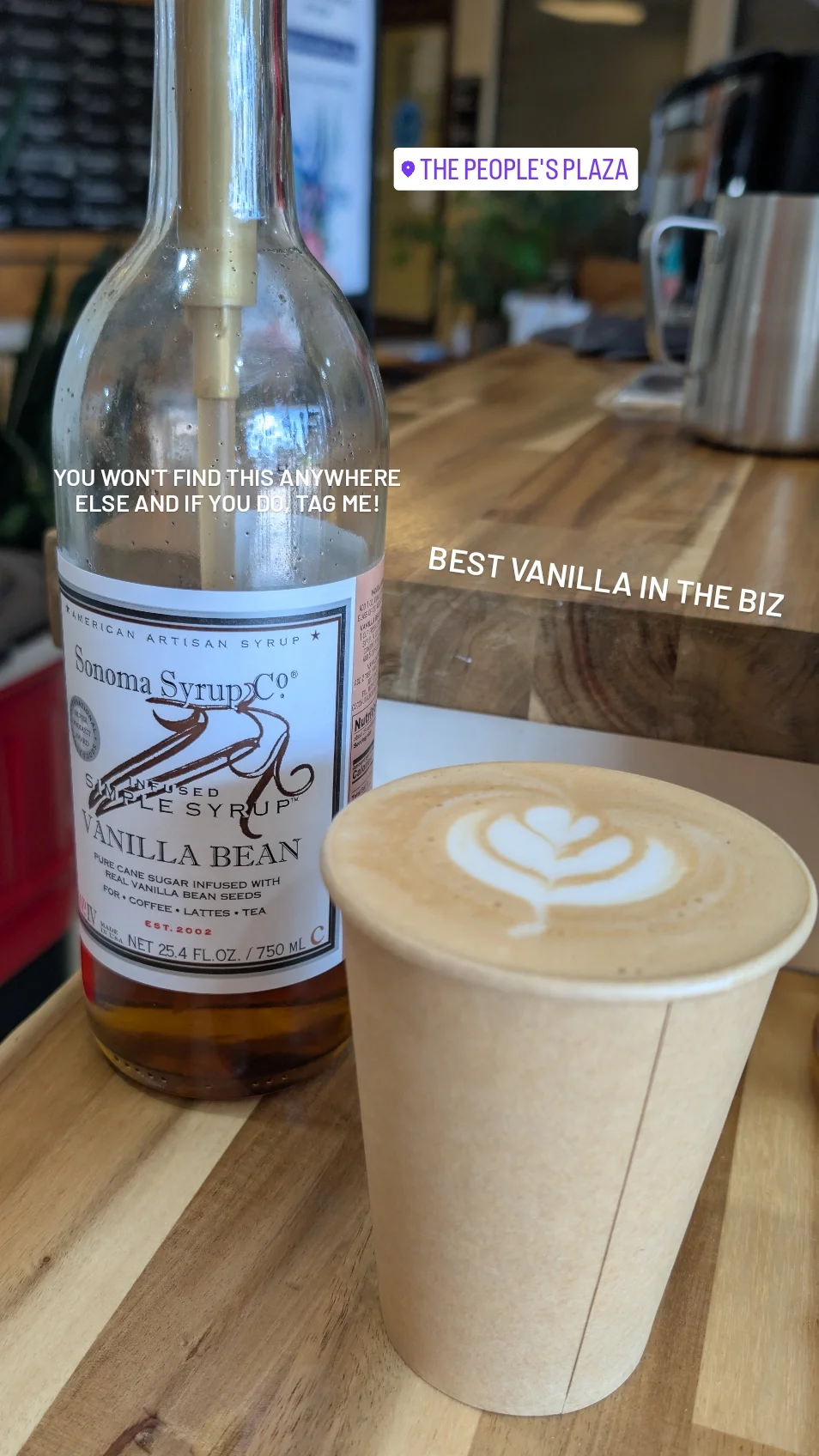 A bottle of Sonoma Syrup Co. vanilla bean syrup and a cup of coffee with latte art on top, sitting on a wooden table in a cafe.