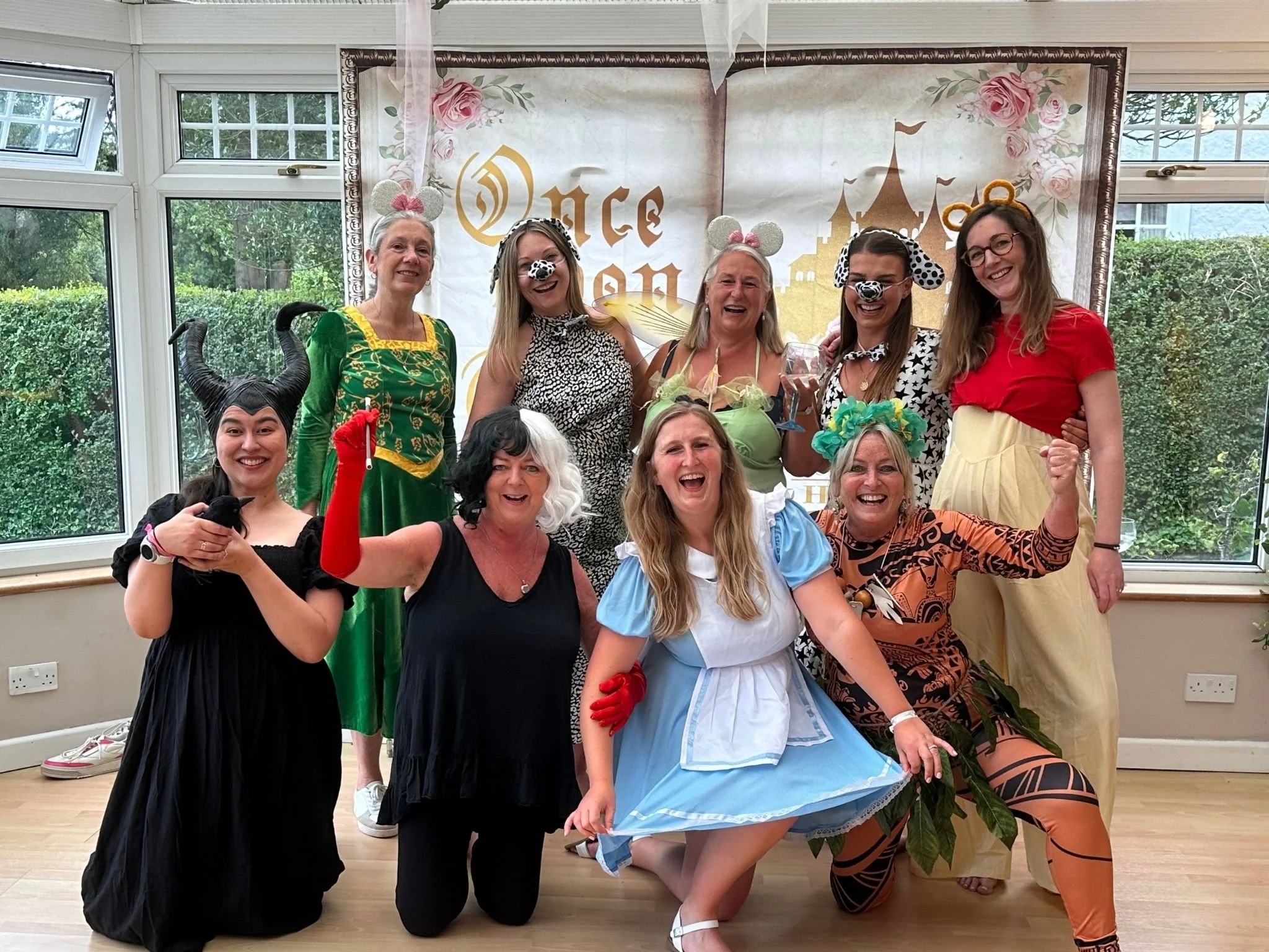 Group of women dressed in animal and fantasy costumes celebrating at a children's party with a 'Once Upon a' themed backdrop, some wearing mouse ears, cow, zebra, and witch costumes, smiling and posing for the photo.
