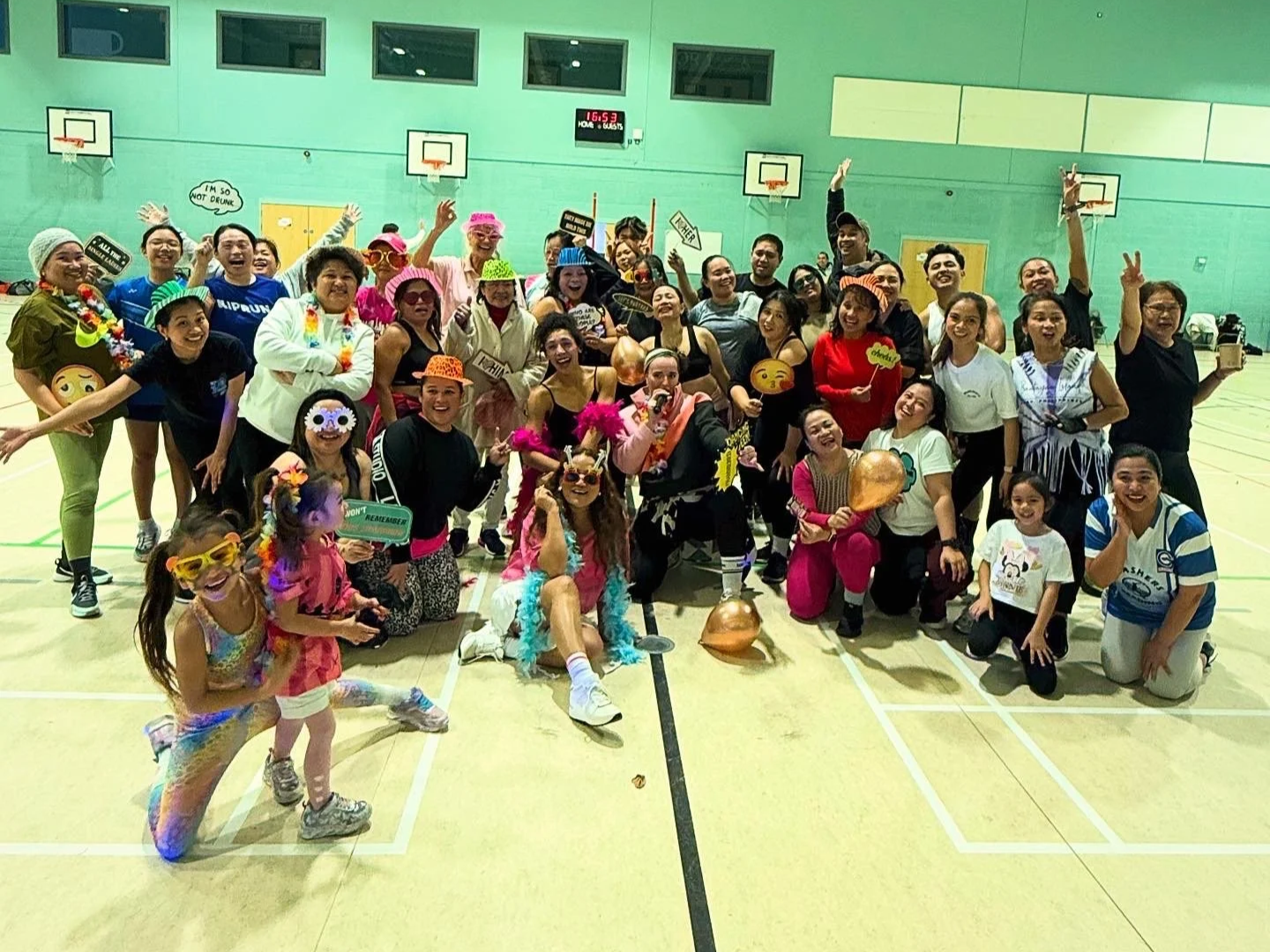 Group of people dressed in colorful costumes and accessories inside a gymnasium, celebrating and holding props, with a basketball hoop and scoreboard in the background.
