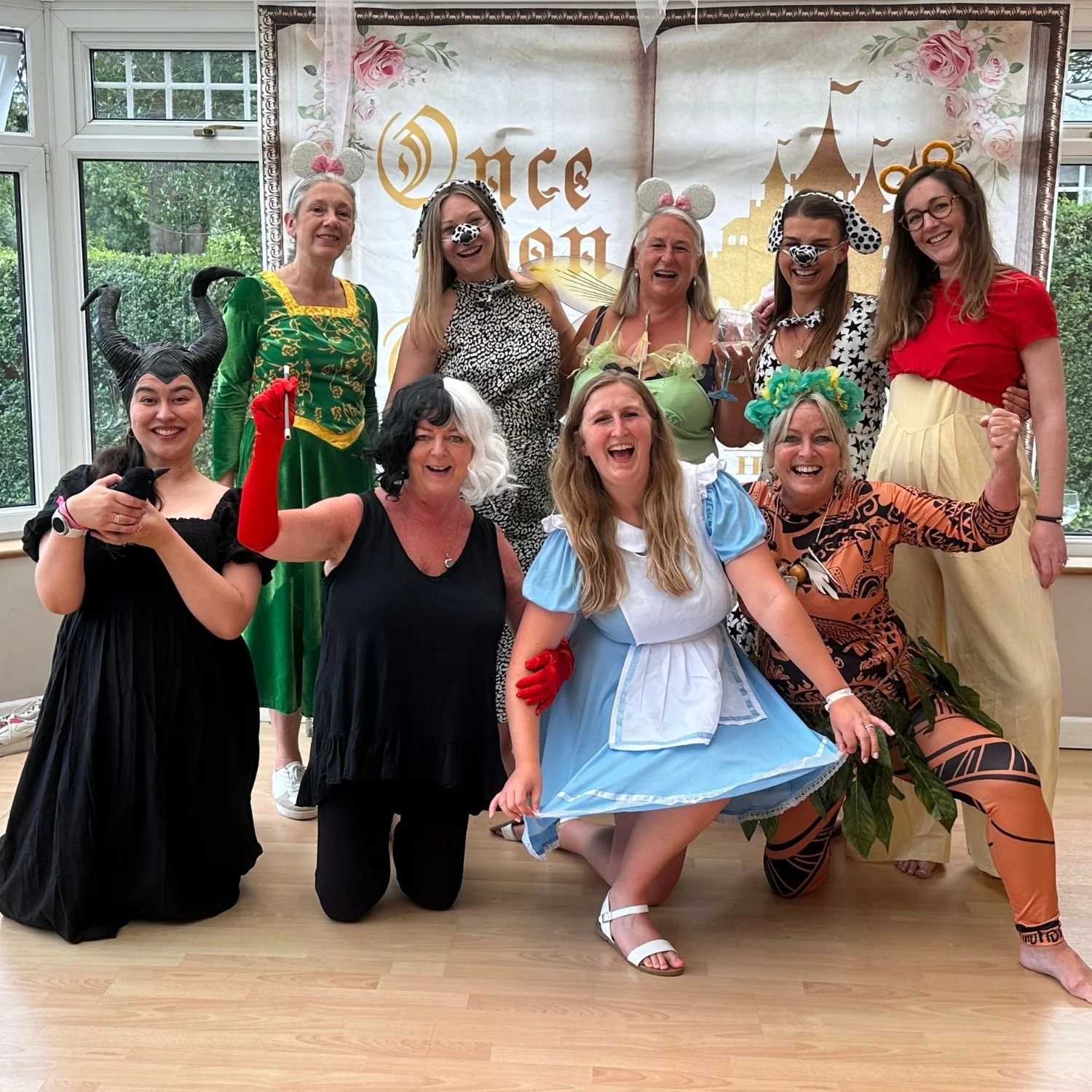 A group of women dressed in costumes from different fairy tale characters and animals, posing happily together at a themed party with a 'Once Upon a Time' backdrop.