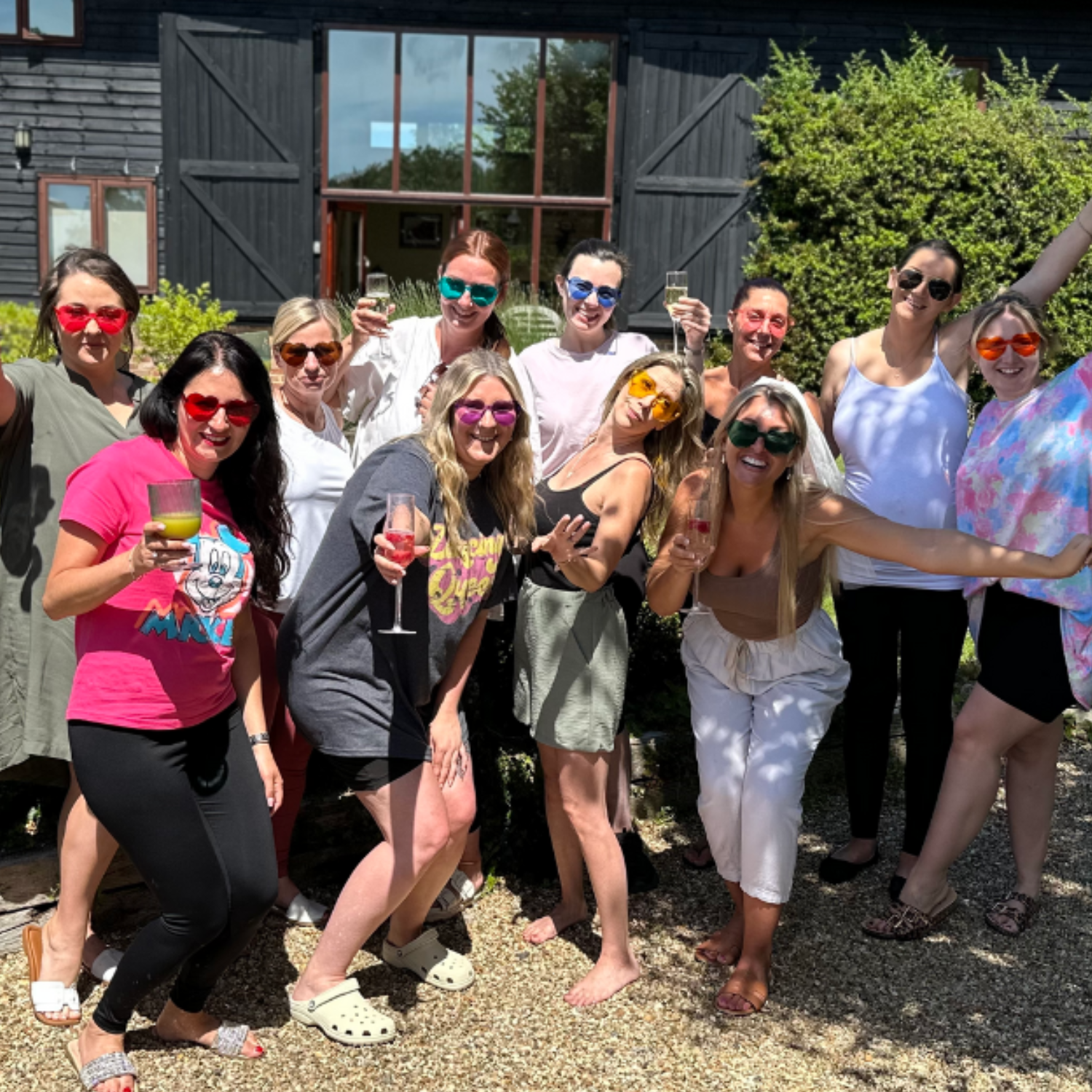 A group of women celebrating outdoors in front of a barn or house with black walls, some holding drinks, all wearing colorful sunglasses and smiling.