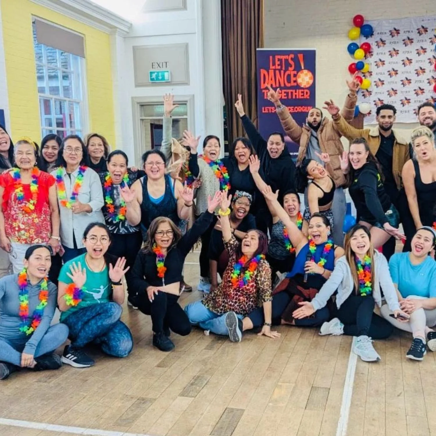 A group of people celebrating and smiling together in a room decorated with colorful balloons and a sign that says "Let's Dance." They are wearing colorful leis, some are raising their hands, and some are sitting or kneeling on the floor.