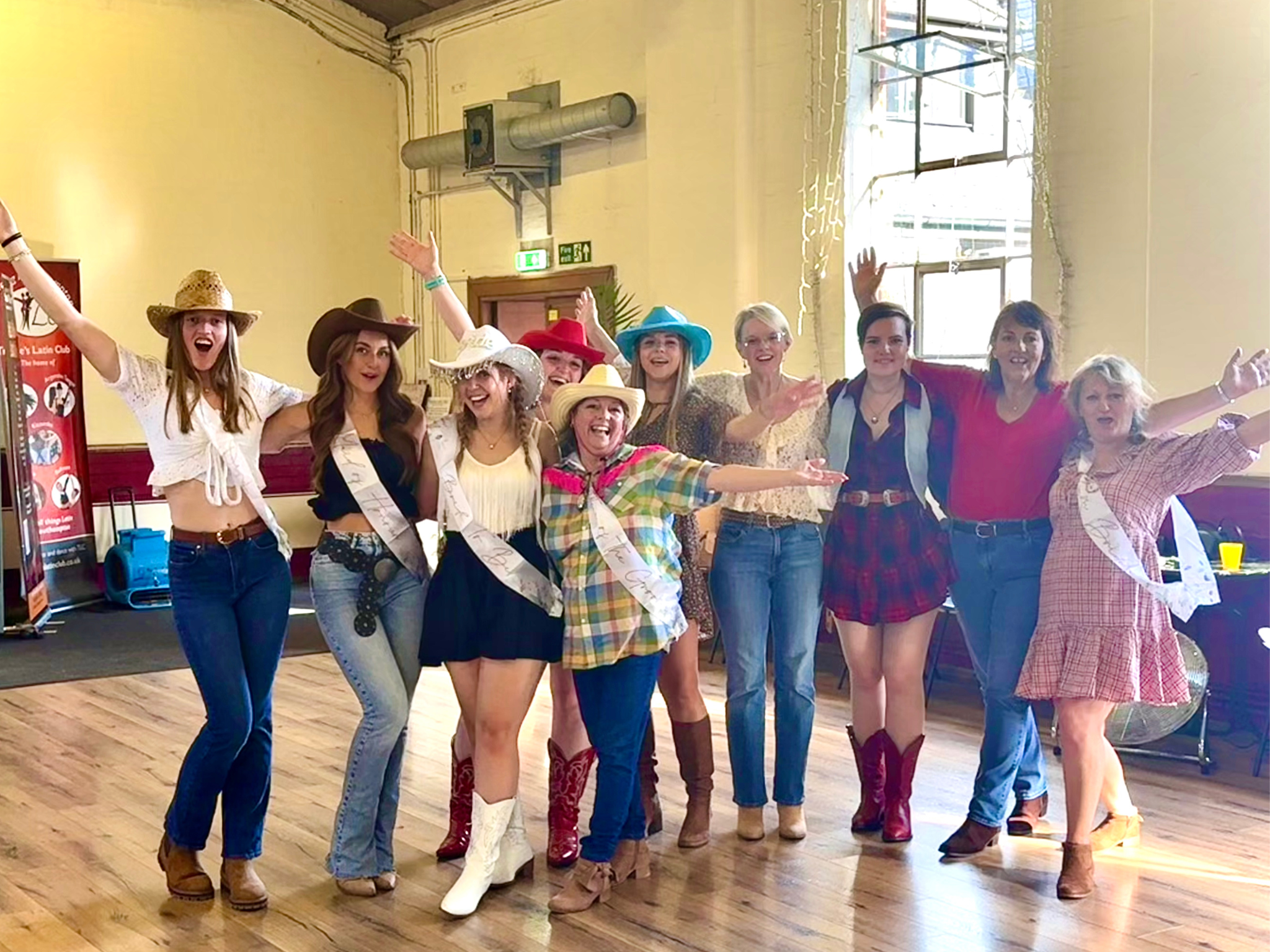 A group of women dressed in country or western-themed outfits, wearing cowboy hats and boots, celebrating together with arms raised and smiling inside a decorated room.