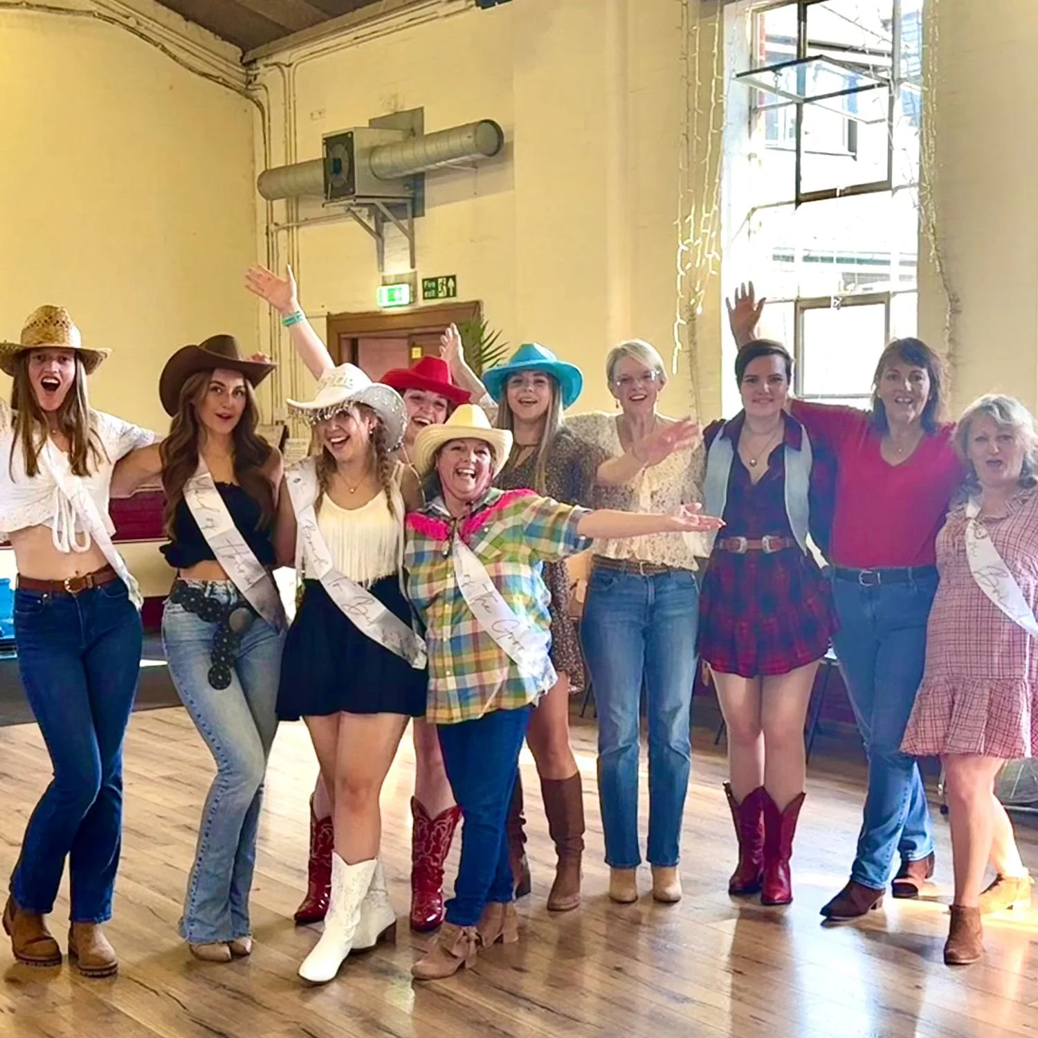 Group of women in cowboy hats celebrating indoors, some with sashes, smiling and raising hands, in a rustic room with wooden floors and large windows.
