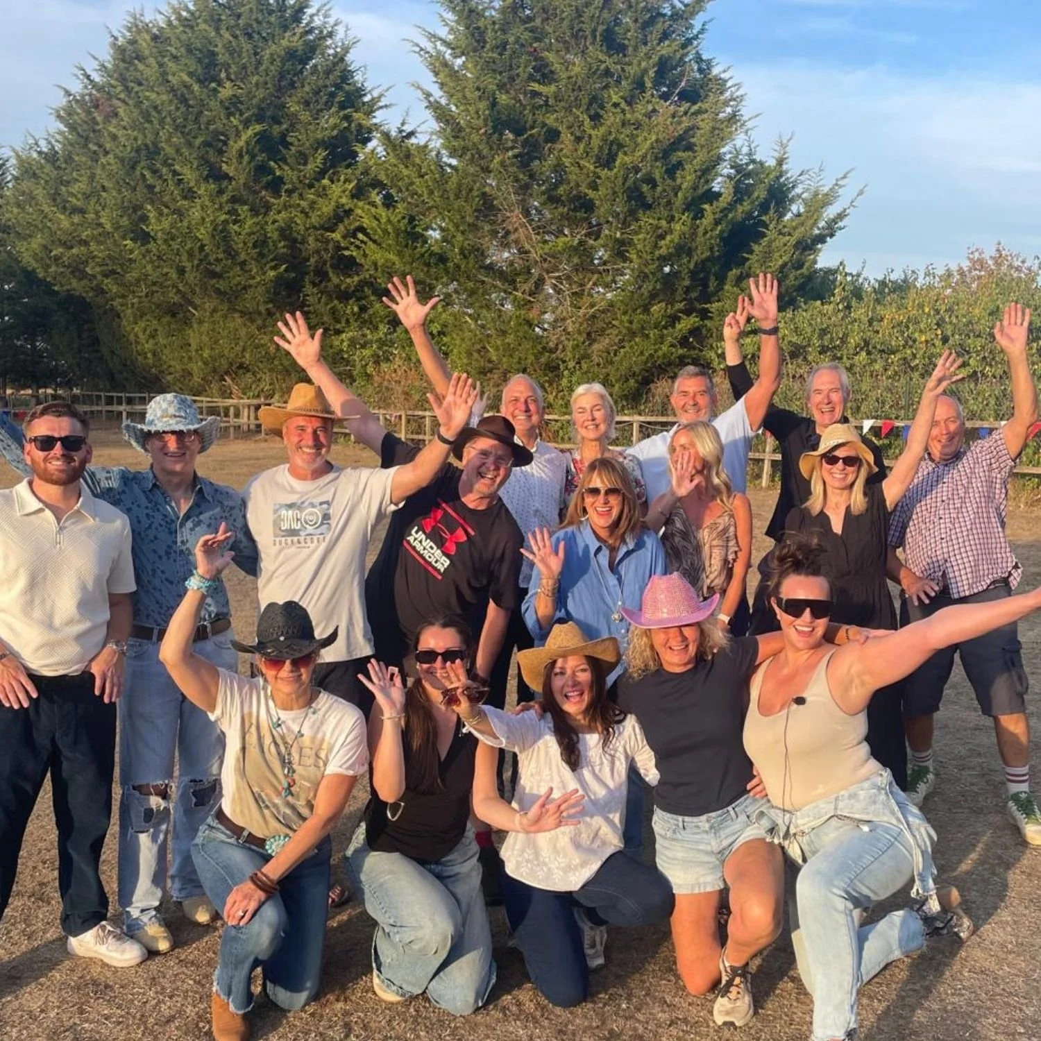 Group of people celebrating outdoors, wearing hats and sunglasses, with trees and a fence in the background.