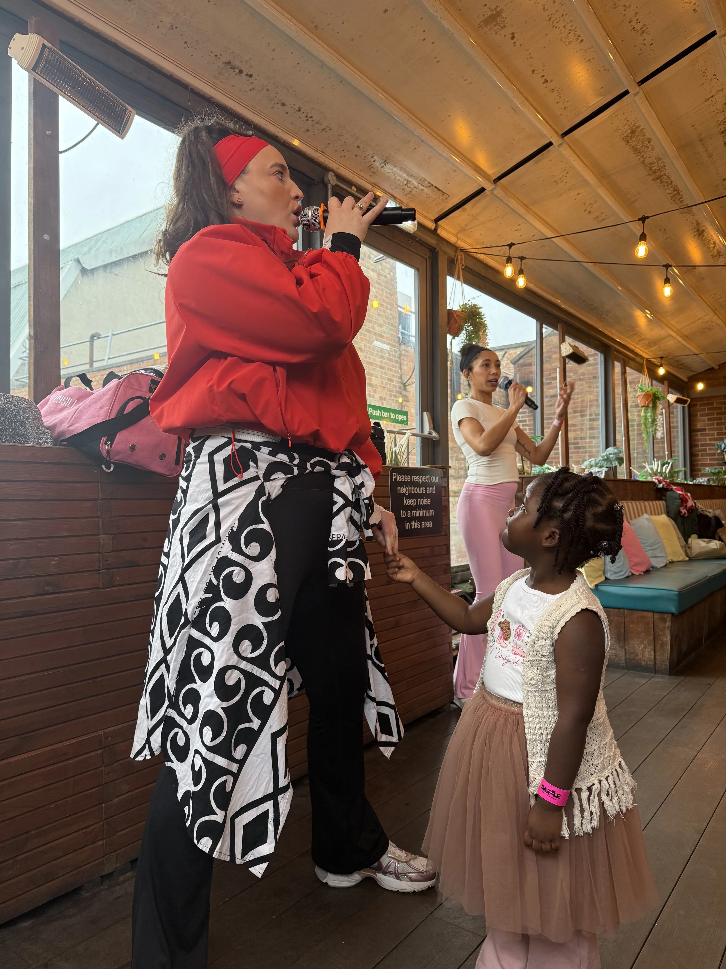 A woman and a young girl are in a cozy indoor space. The woman has long dark hair, is wearing a red jacket, black pants, and a black and white patterned shirt tied around her waist, and is holding a microphone. The young girl, with braided hair, is w
