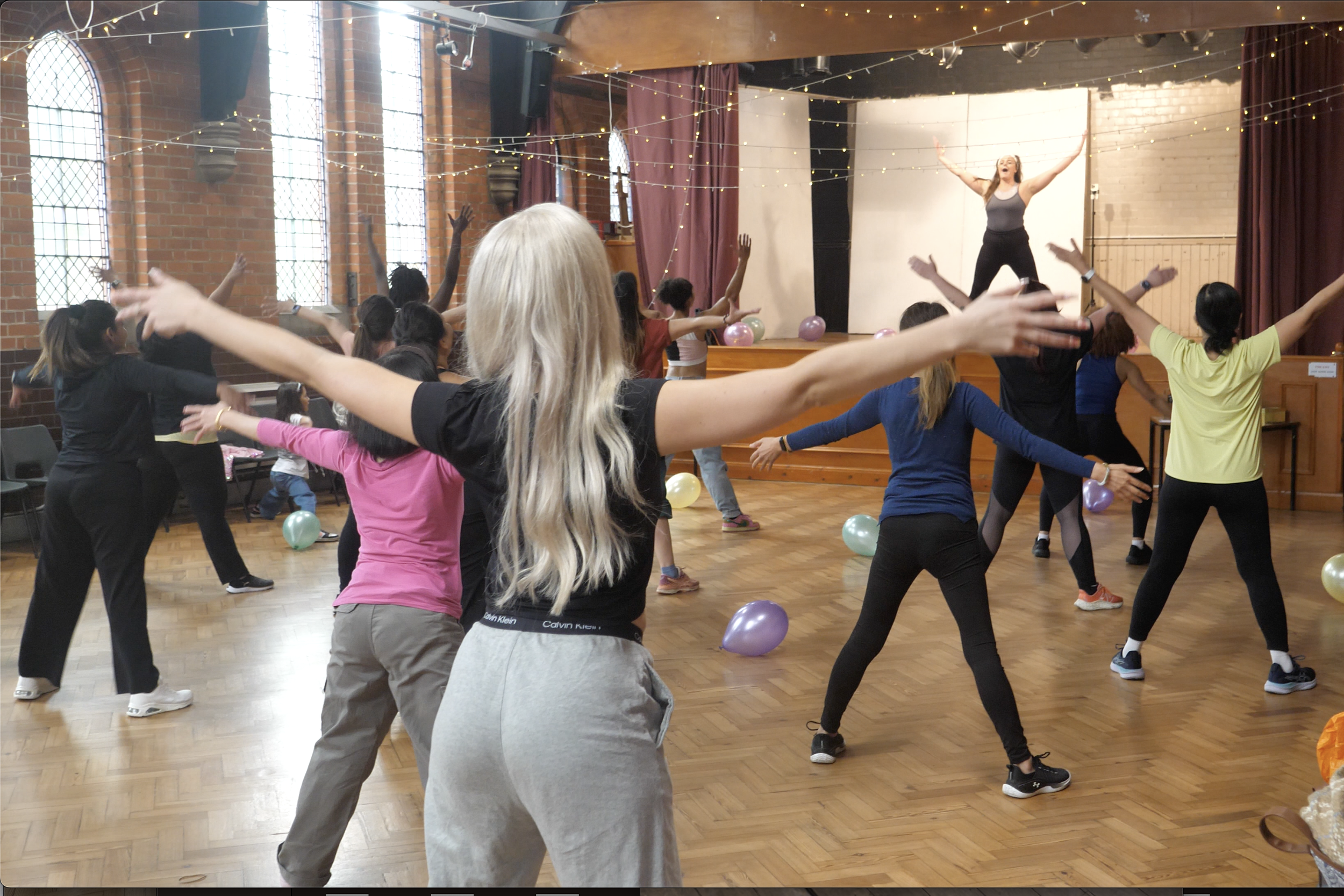 A group of people participating in a dance or exercise class in a spacious room with wooden floors and large windows. The instructor is on a stage at the front, leading the class with arms raised. Balloons are scattered on the floor, and fairy lights