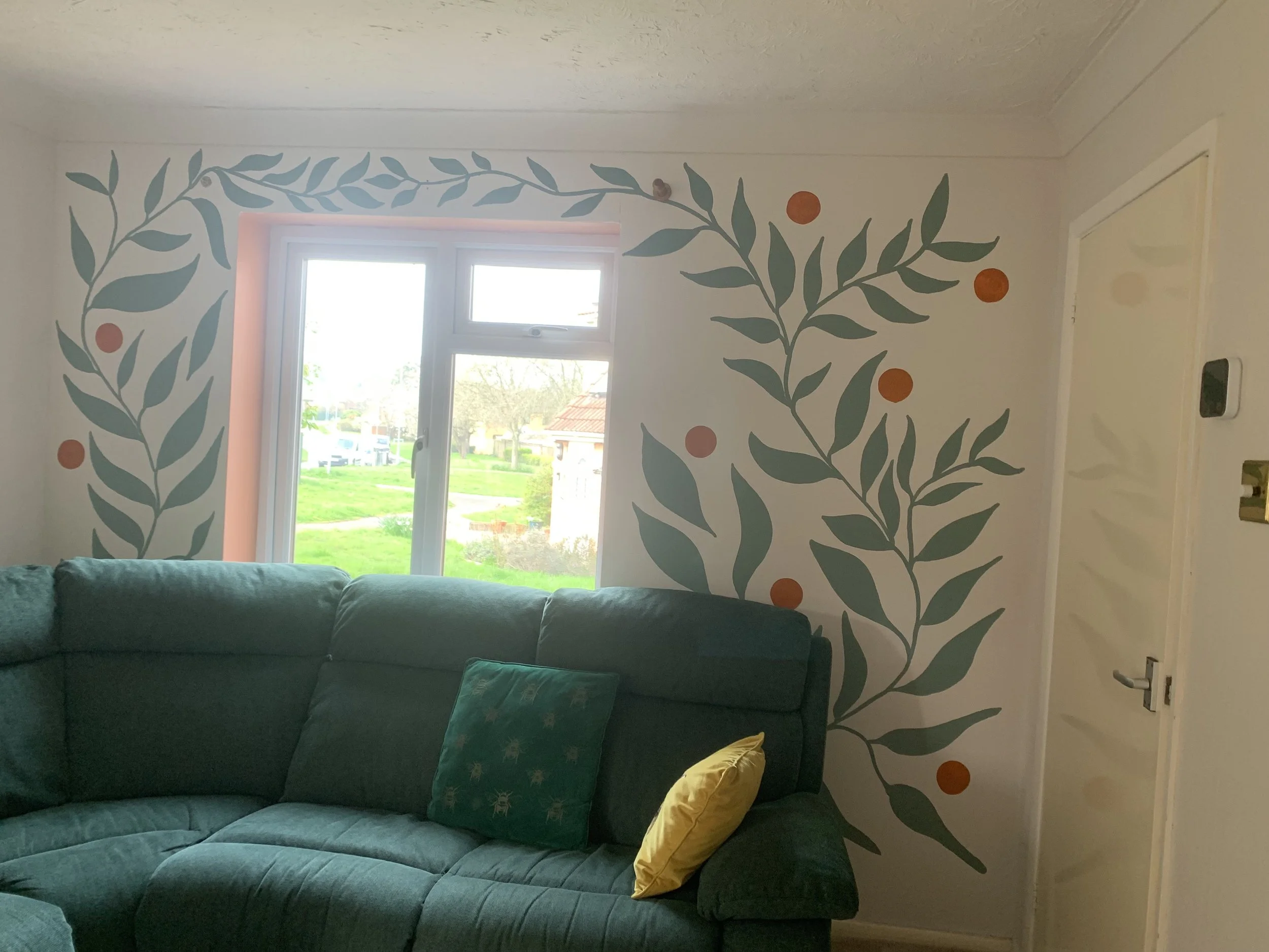 Hand painted mural of green vine leaves and orange circles around a living room window. Green sofa and cushions. Valspar and Dulux paint.