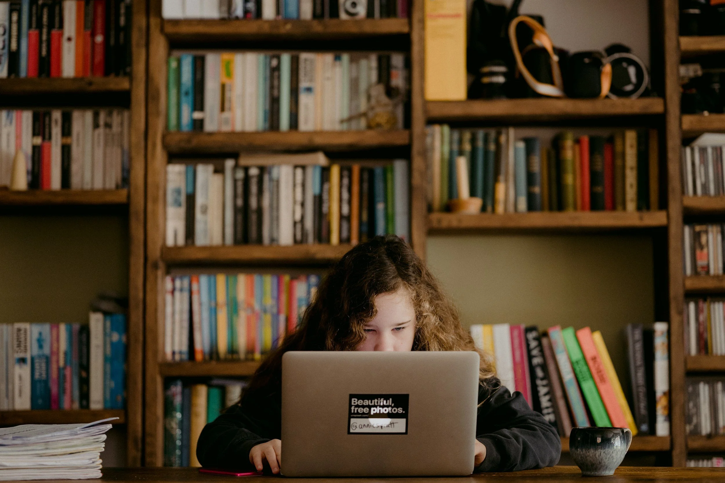 A girl with curly hair working on a laptop at a wooden table in front of bookshelves filled with books.