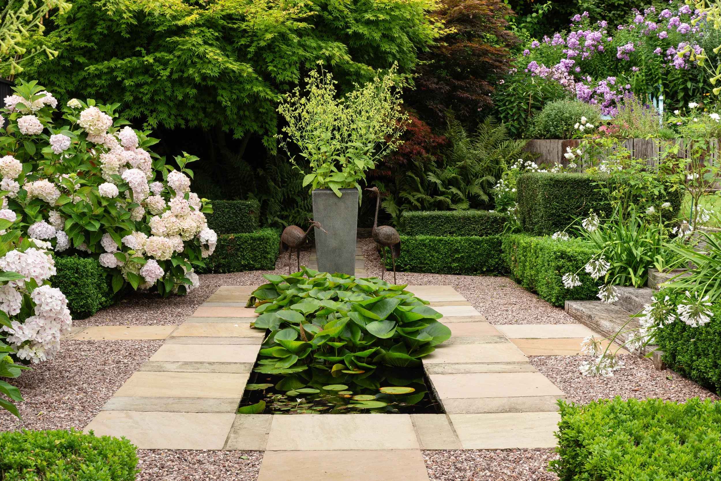 A long rectangular water feature surrounded by flag stone paving, box hedging and white Hydrangeas