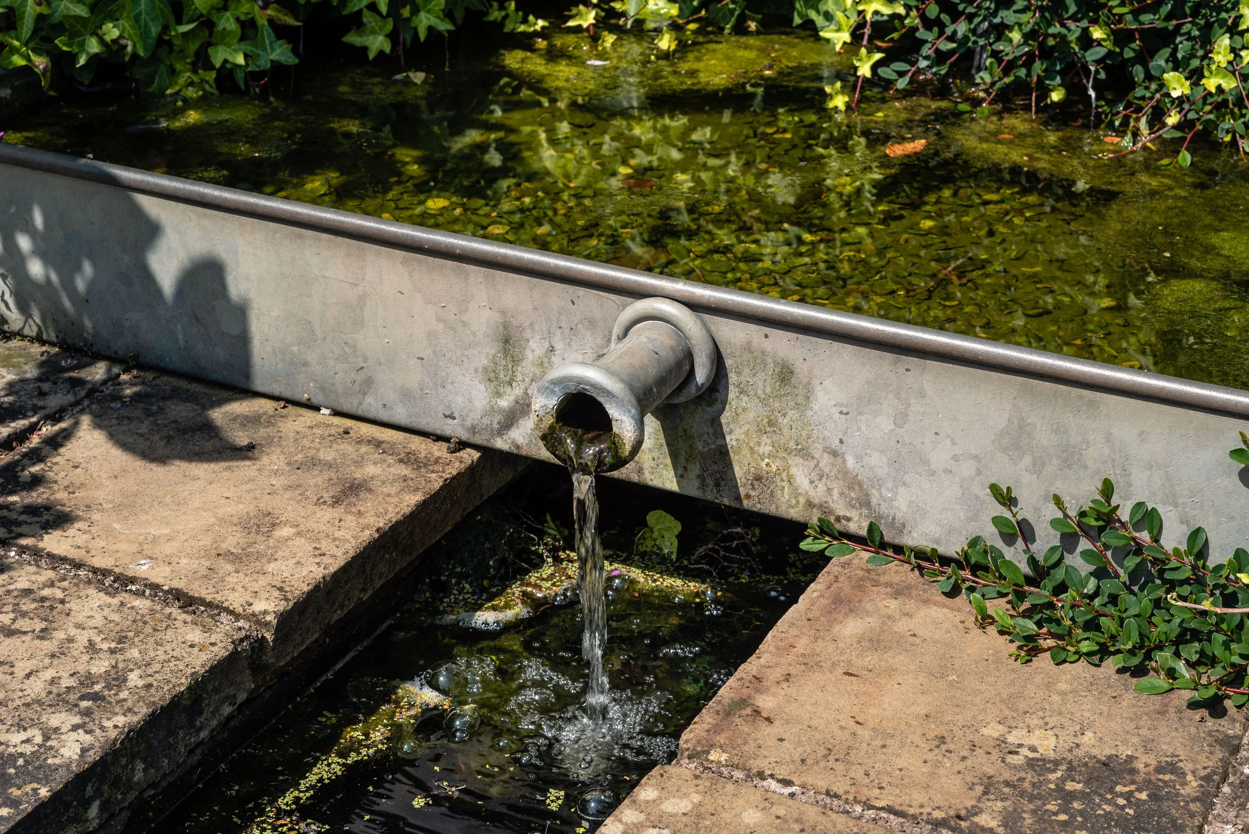 Corton steel water feature with a small stream of water flowing from a pipe into a paving lined rill.