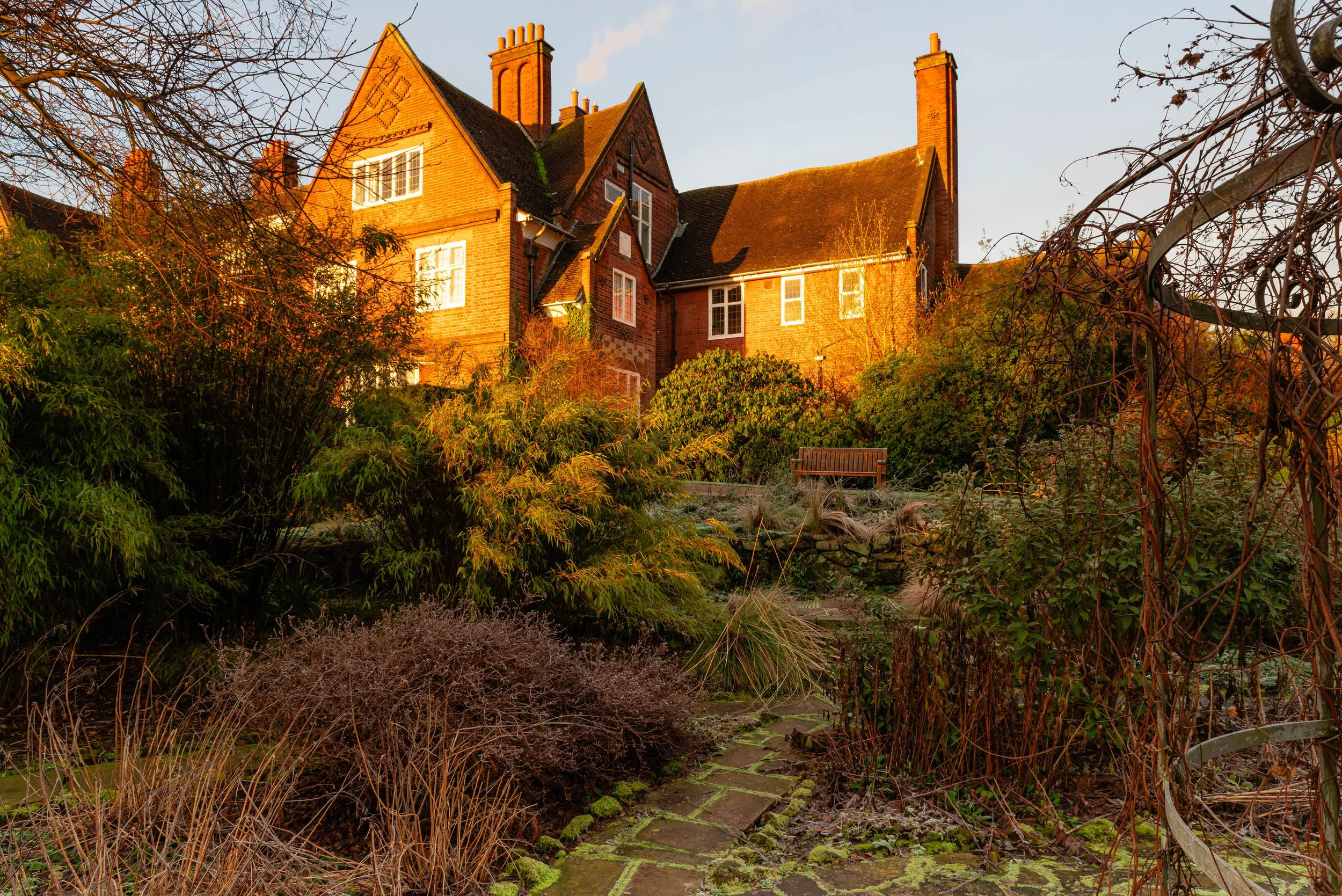 A view up from a shaded, sunken garden, through frosty winter grasses and bamboo, towards a Victorian red-brick mansion bathed in warm early morning sunlight.