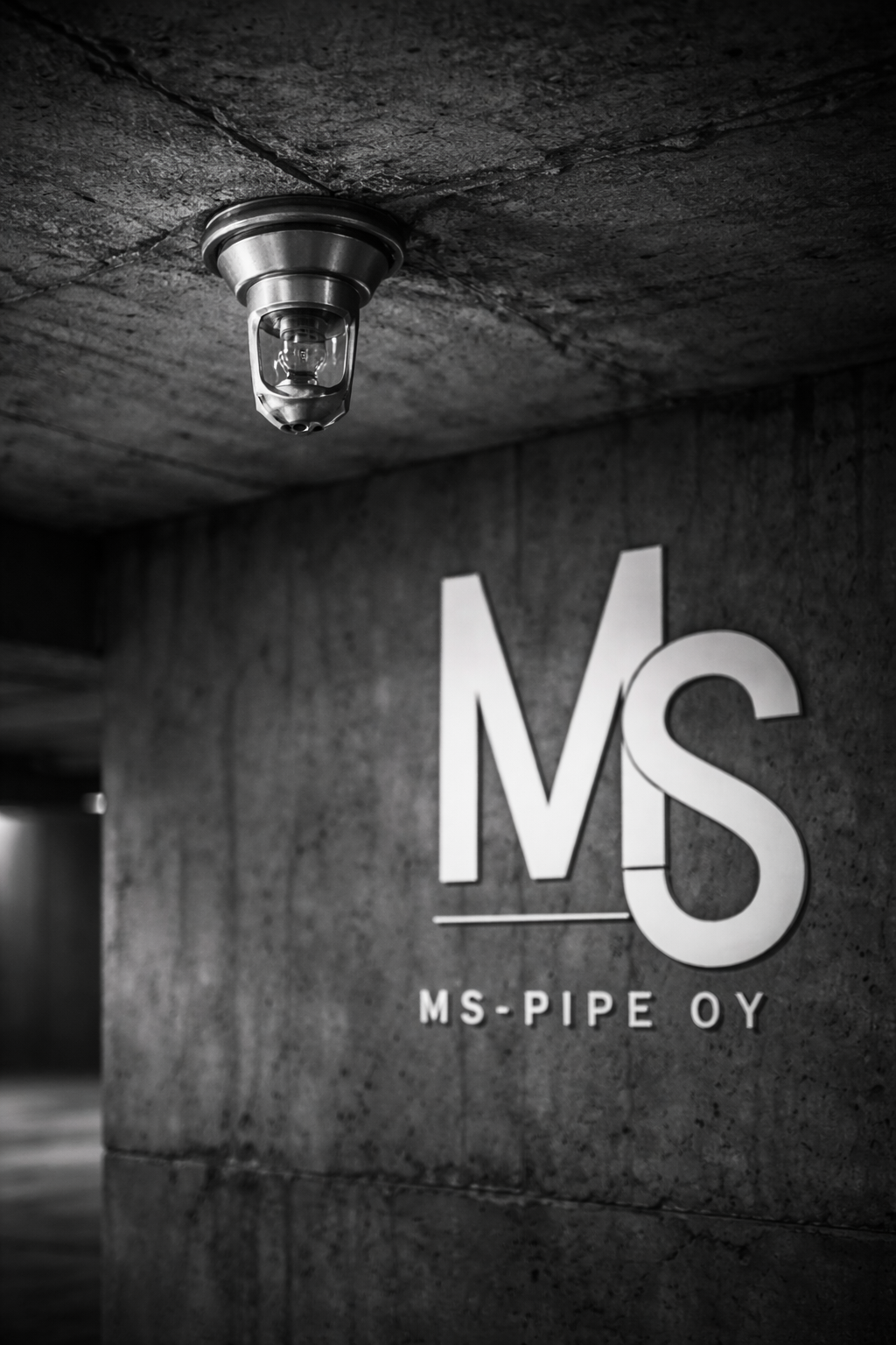 Mustavalkoinen kuva sisätilasta, jossa näkyy ISO-valaisin katossa ja suuret valkoiset kirjaimet 'MS' seinällä. Alla teksti 'MS-PIPE OY'.