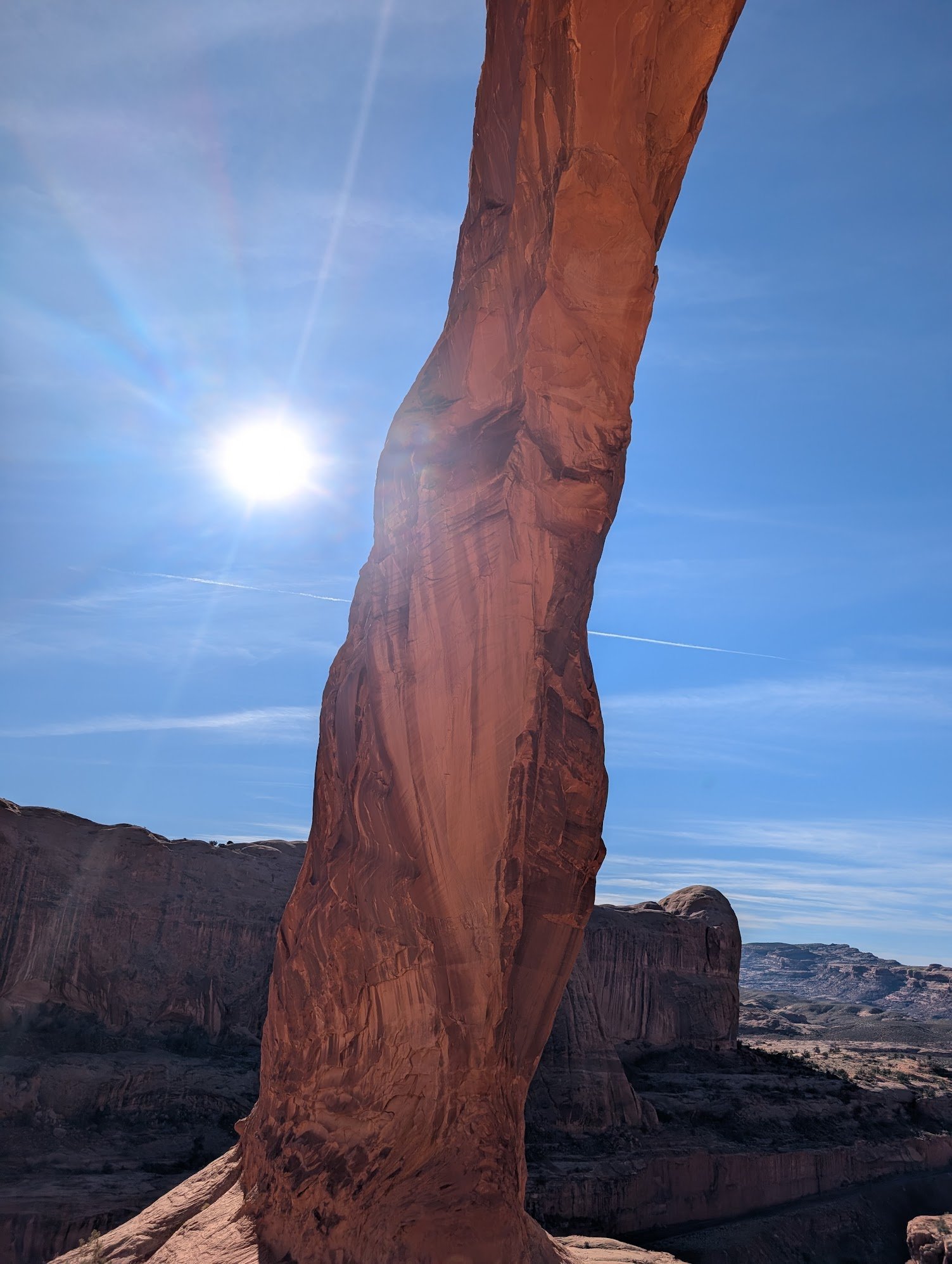 A tall, narrow sandstone formation in a desert landscape with clear blue sky and bright sun