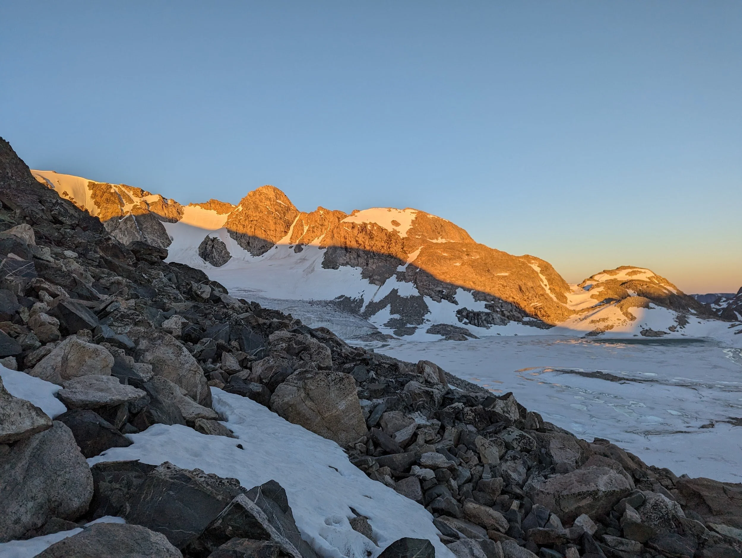 Mountain landscape with rocky foreground, snow, and glaciers, illuminated by the setting or rising sun under a clear blue sky.