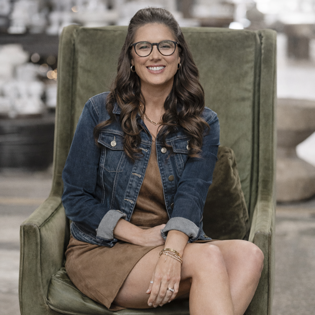 Moss Artist Lindsay Johnson with long brown hair wearing glasses, a denim jacket, and a brown dress sitting on a green armchair, smiling in an indoor setting.