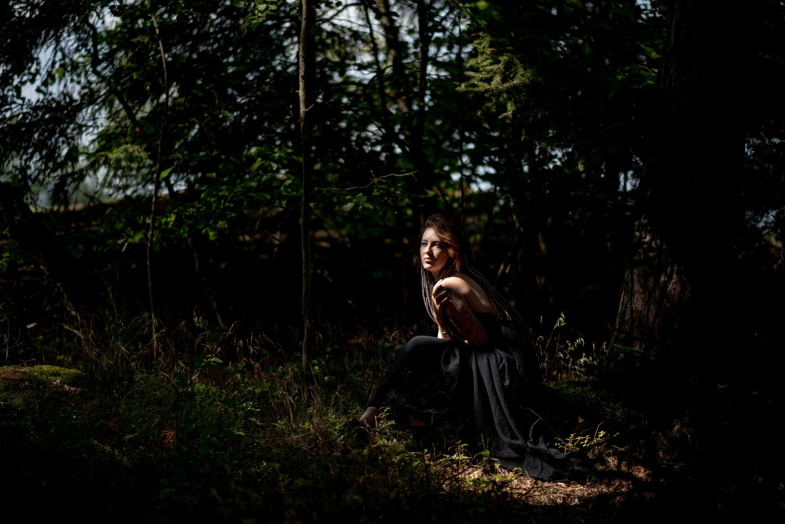 A woman with tattoos sitting in a forest, illuminated by sunlight amidst dark trees and foliage.