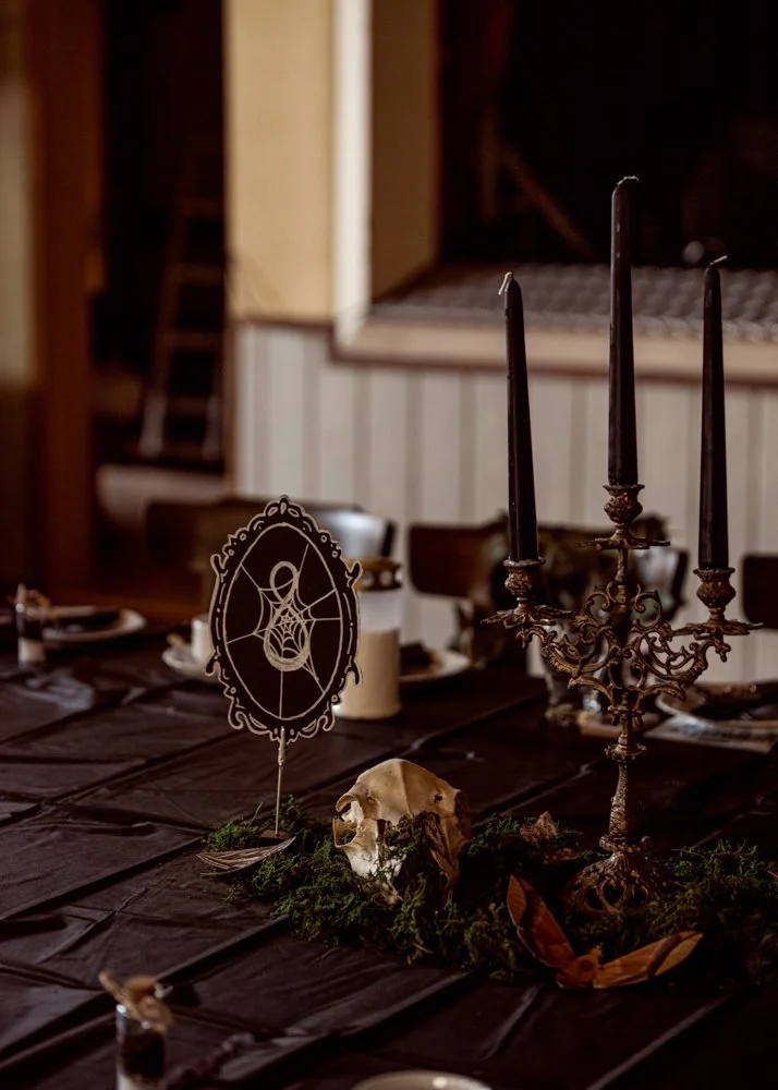 A dark-themed table setting with a skull, a decorative black and gold candelabra, lit candles, and a sign with a spider web design, possibly for a Halloween or gothic event.