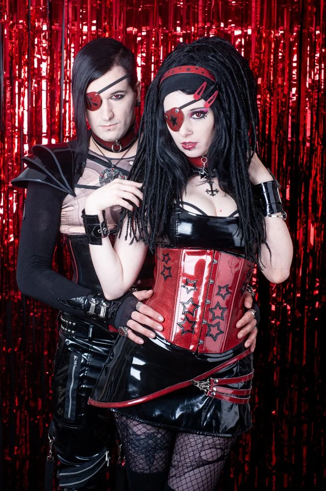 A couple in gothic punk fashion posing in front of a red tinsel background. They wear black and red outfits, including an eye patch and accessories, with dark makeup and styled hair.