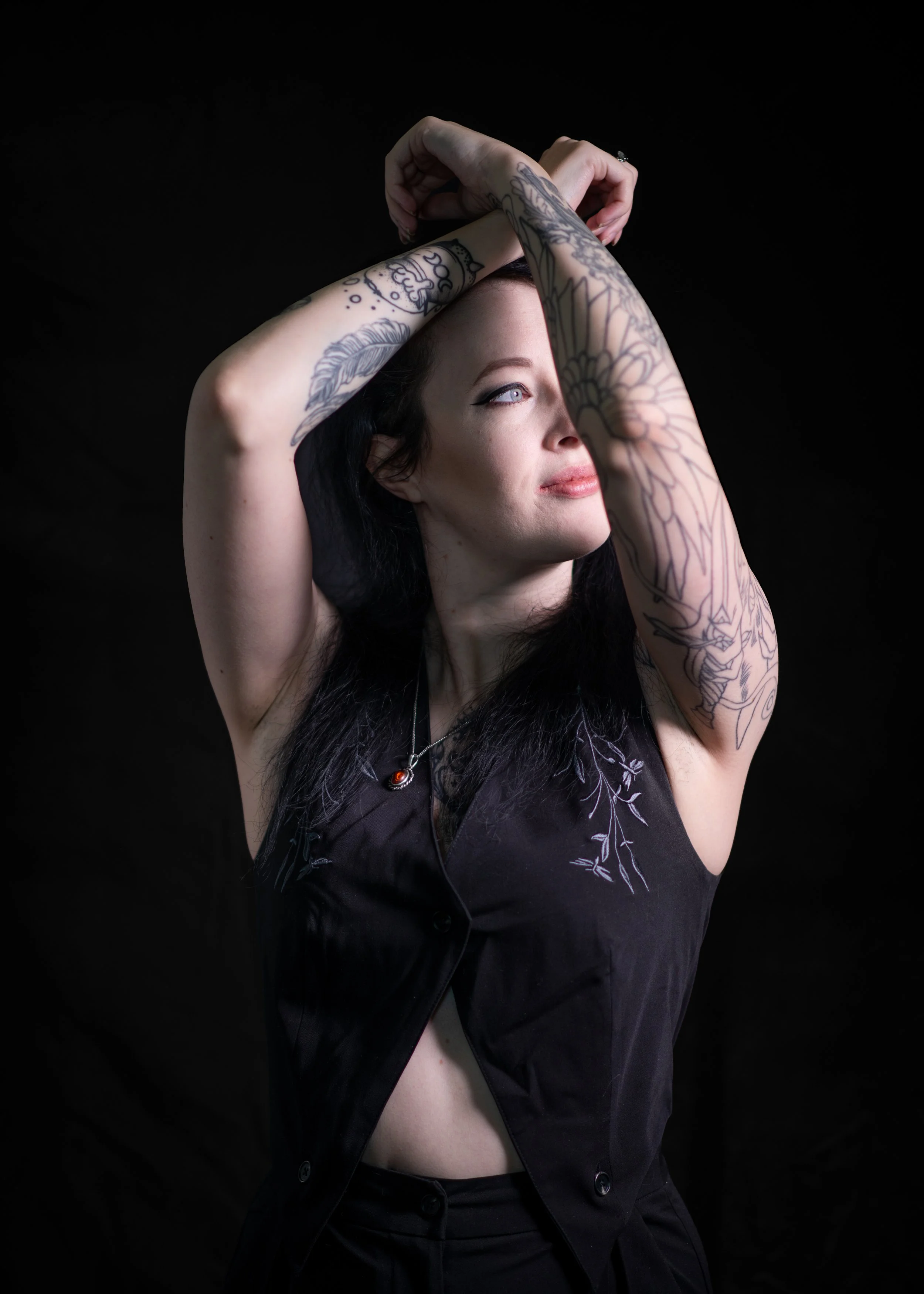 A woman with dark hair and tattoos on her arms, wearing a black sleeveless top, striking a pose with her arms above her head against a black background.