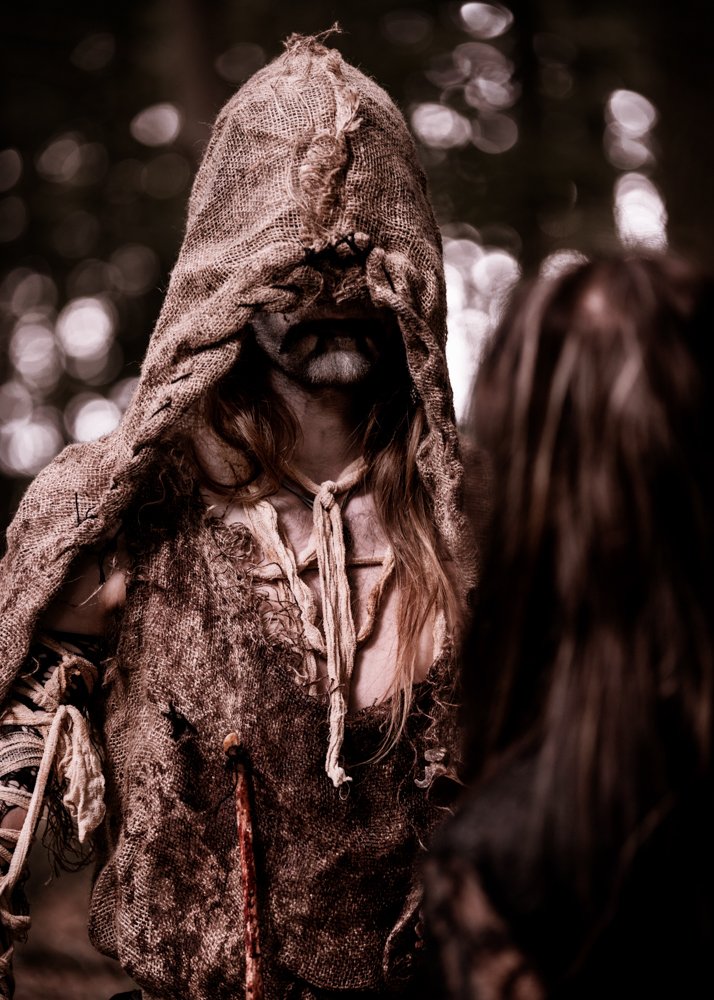 Person dressed in a rustic, tattered costume with a hood and mask made of burlap, resembling a scarecrow or a woods-dwelling character, in a forest setting.
