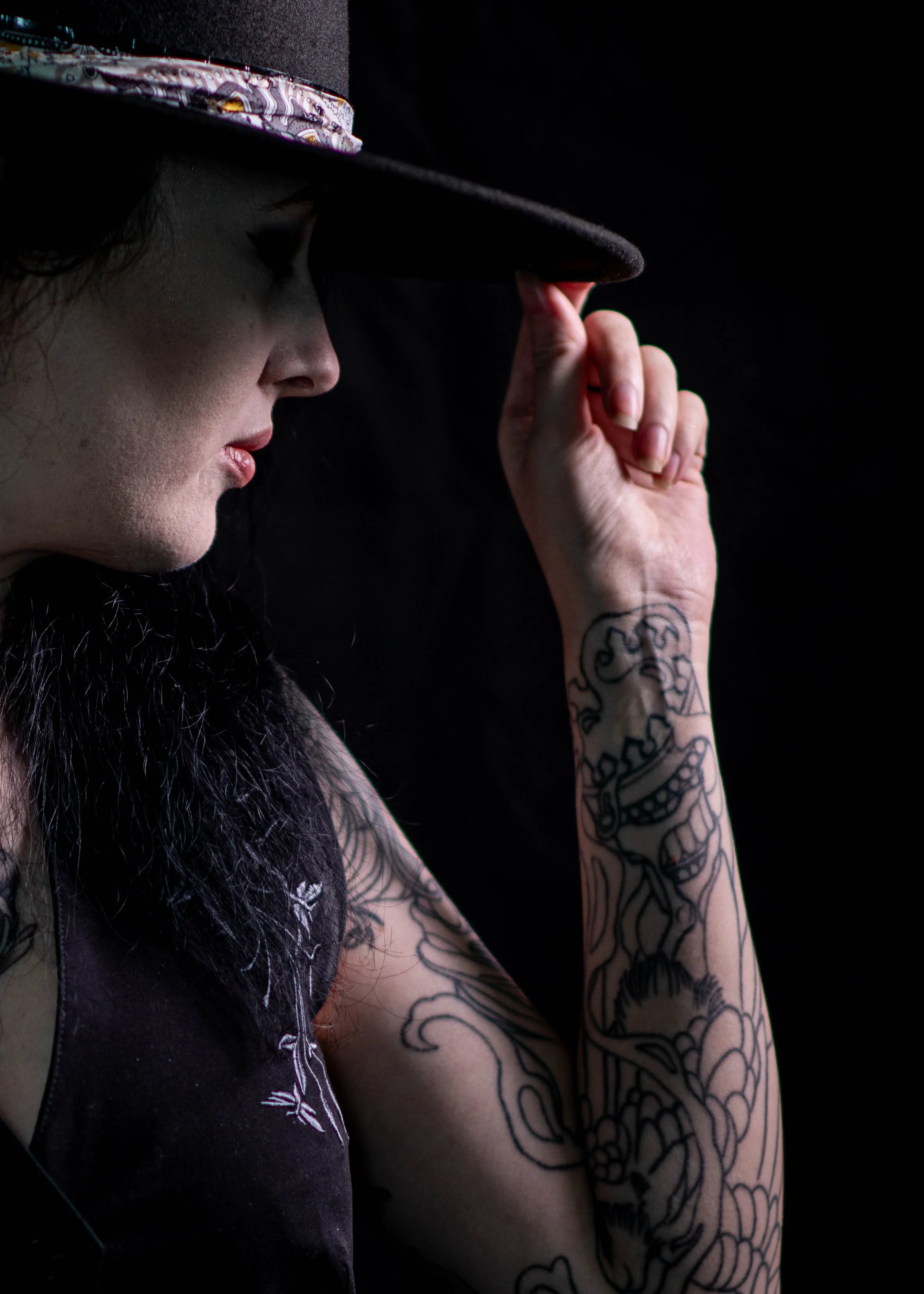 A woman with dark hair and tattoos on her arm, wearing a hat with a floral band, tilting her head downward and holding the brim of her hat against a black background.