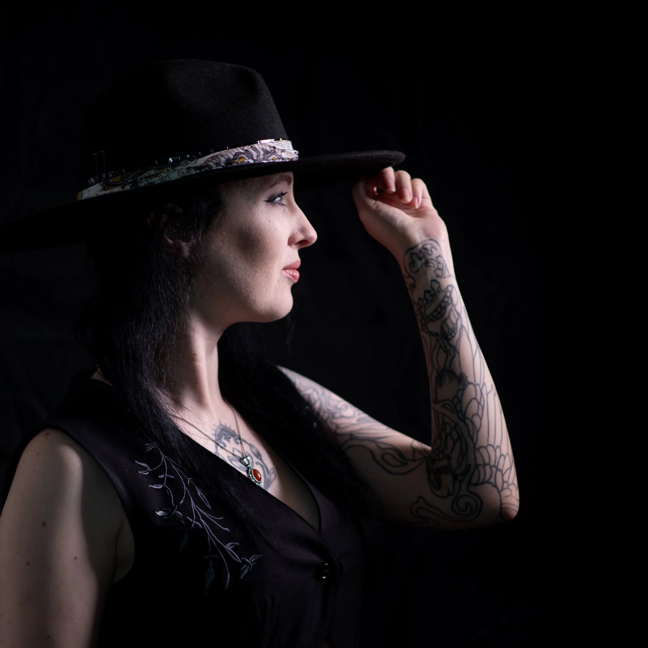 A woman with dark hair and tattoos on her arm, wearing a black sleeveless top and a large black hat with a decorative band, posing against a black background.