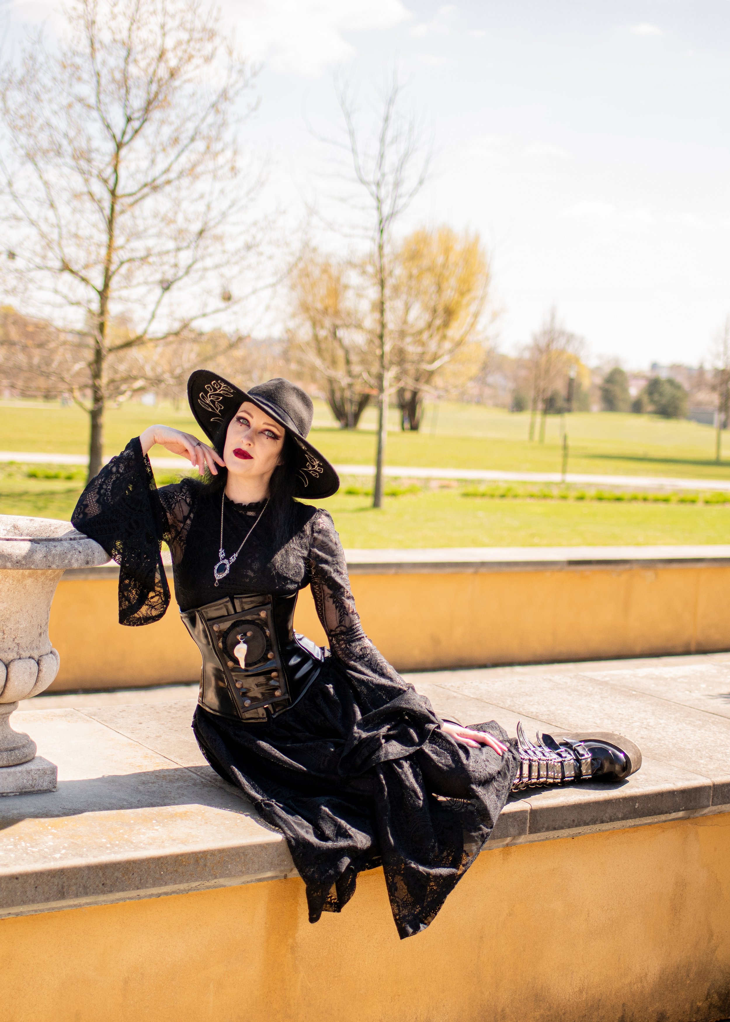 A woman dressed in Gothic style attire with black lace dress, wide-brimmed hat, and corset, sitting on a park bench near a stone ledge on a sunny day with leafless trees in the background.