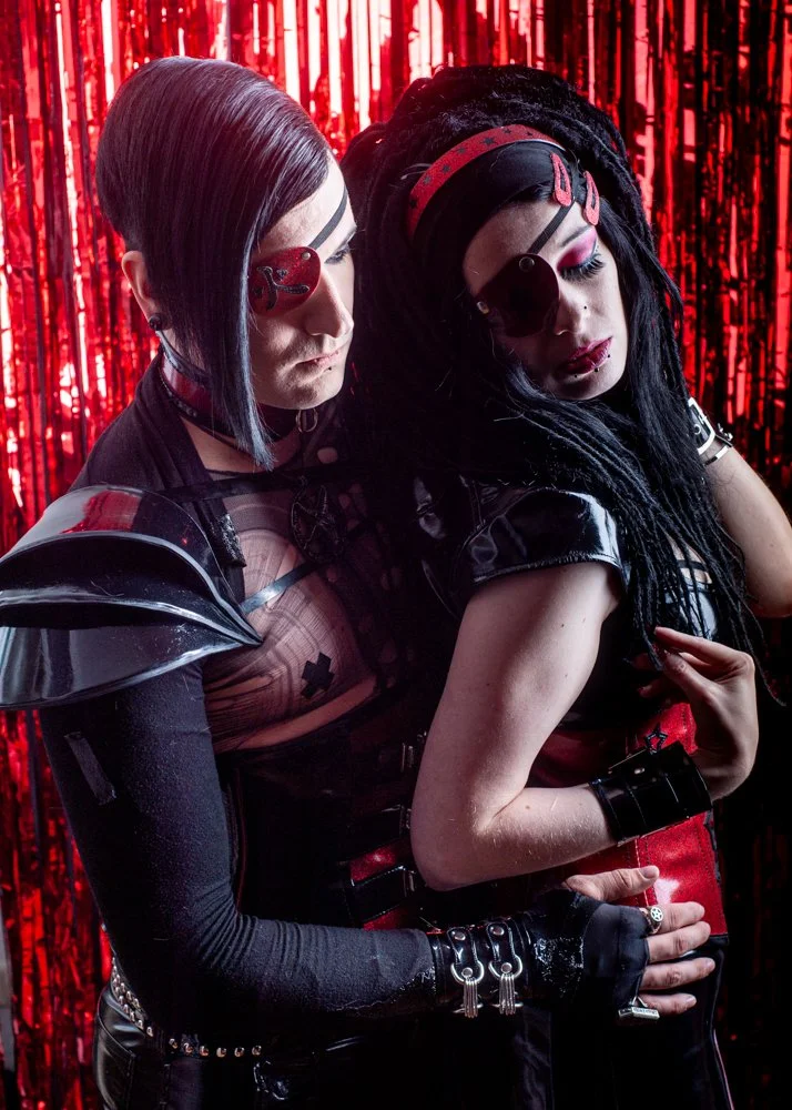 A couple dressed in goth fashion standing close together against a red tinsel background, with one person gently holding the other from behind.