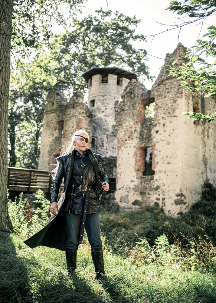 A man dressed in a black leather jacket, pants, and tall boots standing outdoors near an old stone castle surrounded by trees.
