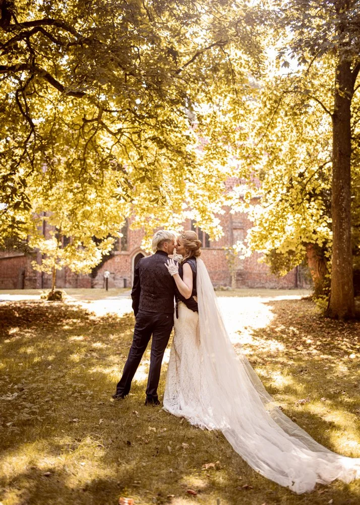 A bride and groom share a kiss outdoors under autumn trees with sunlight filtering through the leaves, in front of an old brick building.