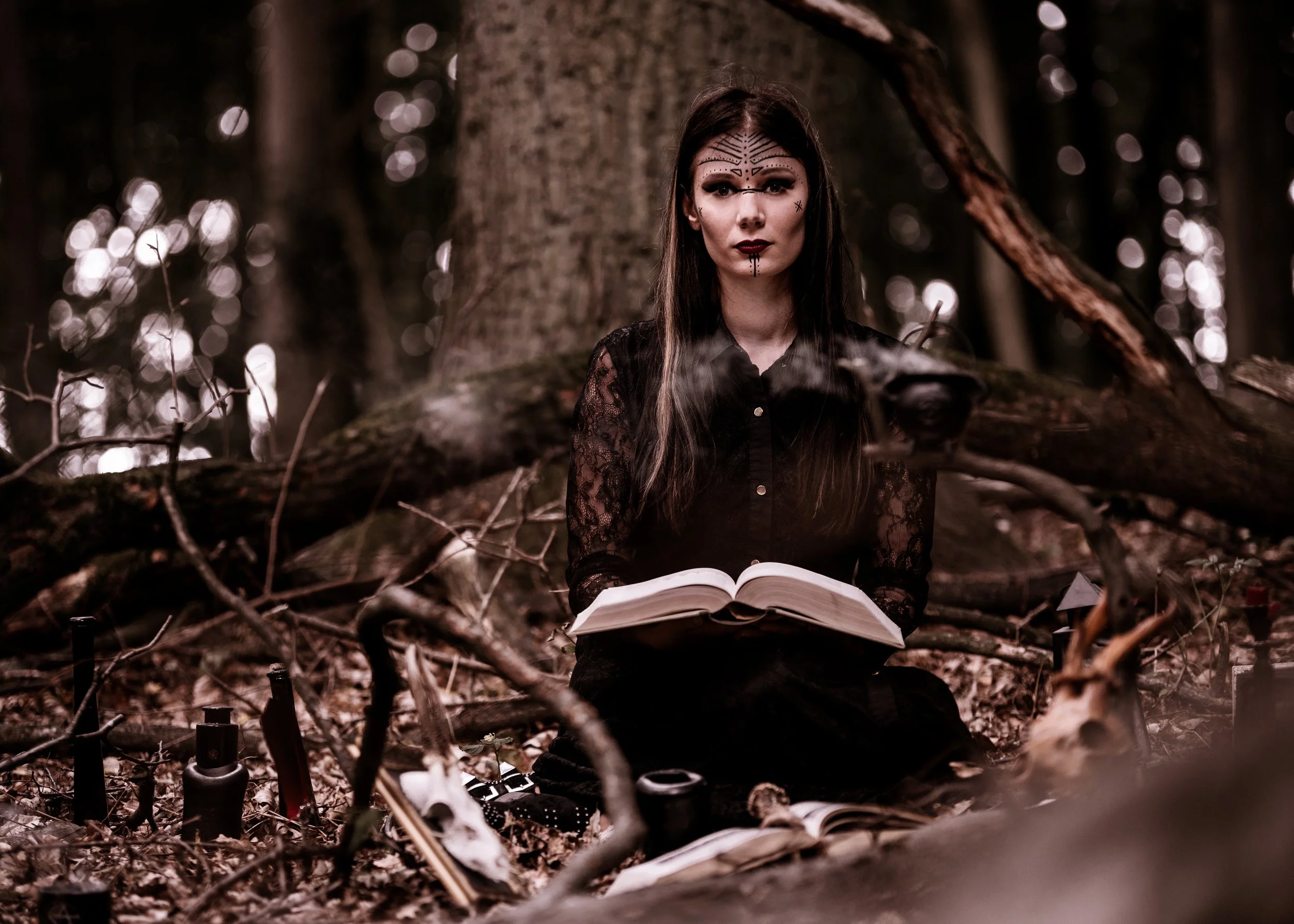A woman with dark hair and face tattoos wearing black lace clothing, sitting in a forest surrounded by fallen branches and skulls, holding an open book with a smoky cup nearby, creating a mystical atmosphere.